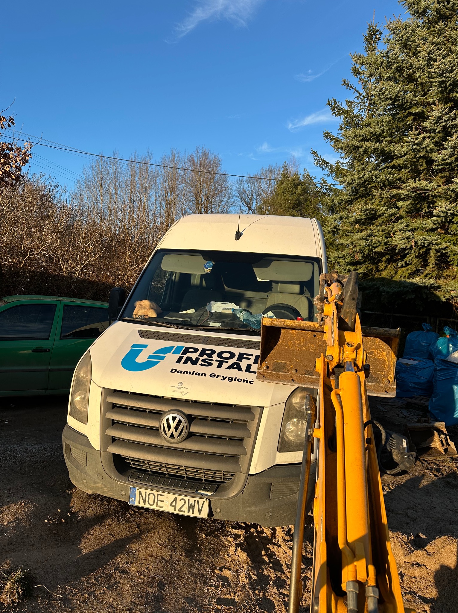 Biały van z logo 'U Profi Instal' i numerem rejestracyjnym NOE 42WV, zaparkowany na placu budowy, z widoczną koparką i workami z odpadami w tle.