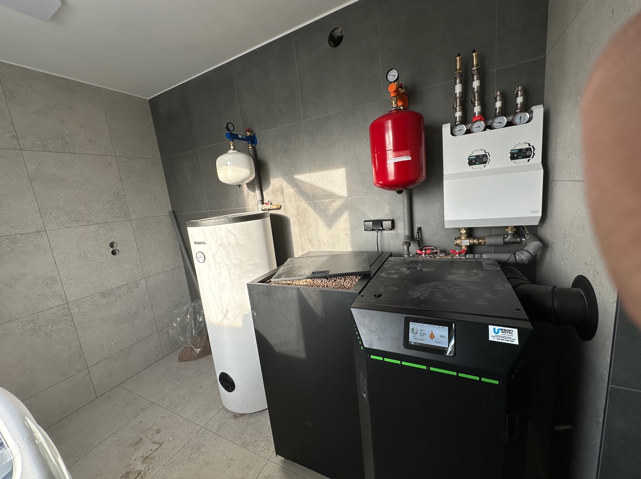 Nowoczesna kotłownia z piecem na pellet, zbiornikiem buforowym i osprzętem. Widoczne elementy instalacji grzewczej w pomieszczeniu wykończonym płytkami.