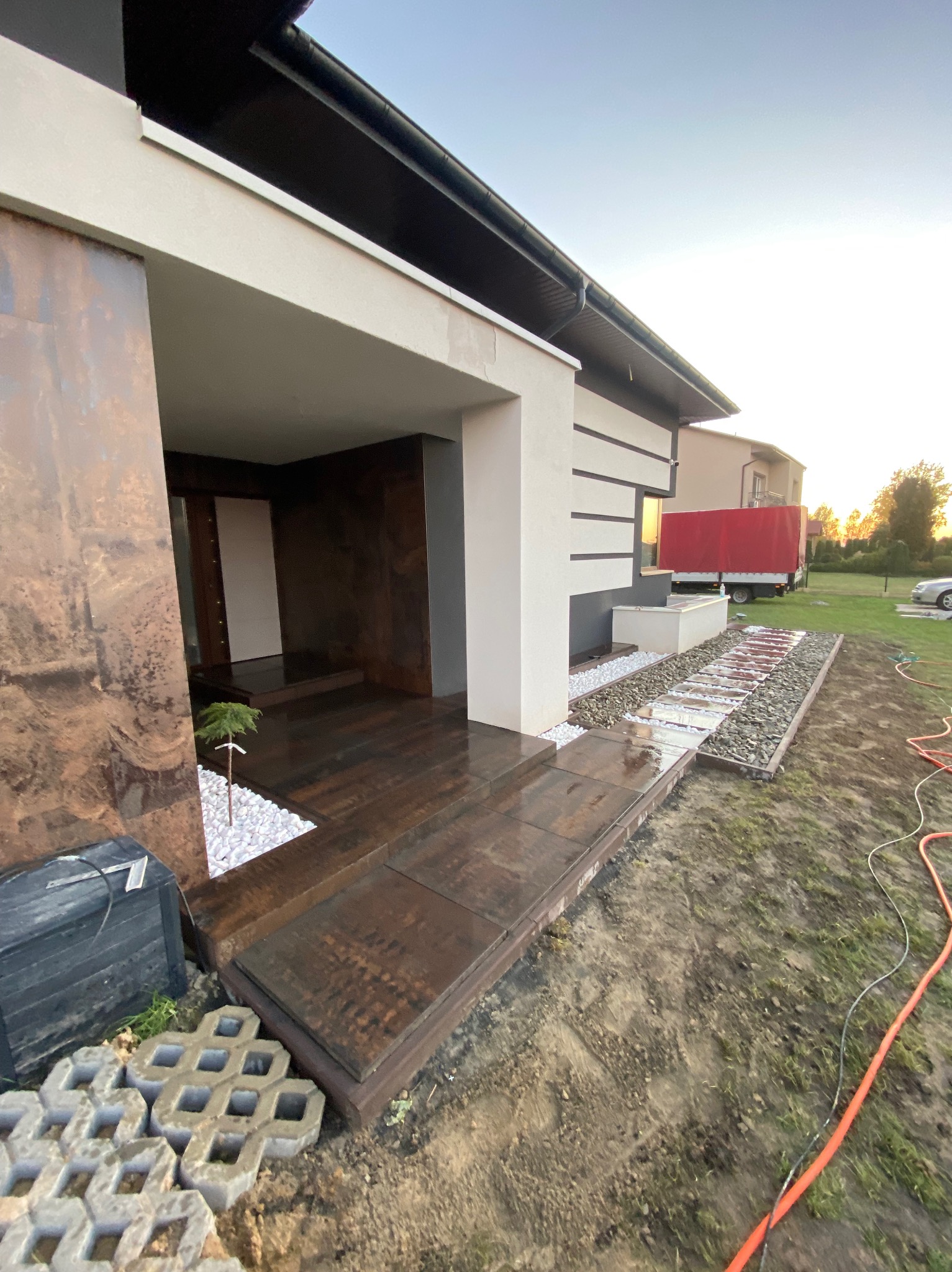 Nowoczesny dom jednorodzinny z oryginalnym wejściem wykończonym płytami imitującymi rdzę, białymi kamykami i ścieżką z płyt na żwirowym podłożu.