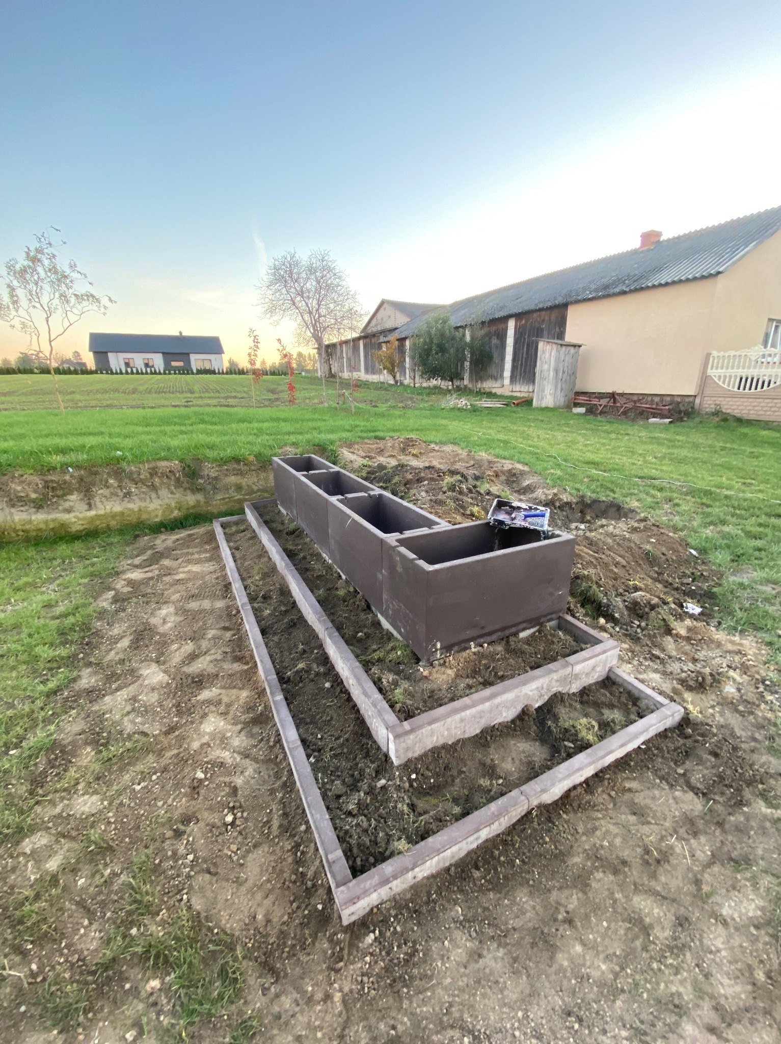 Montaż podwyższonych rabat z betonowych elementów na działce. Widoczne przygotowane podłoże i częściowo złożona konstrukcja. W tle budynki i zieleń.