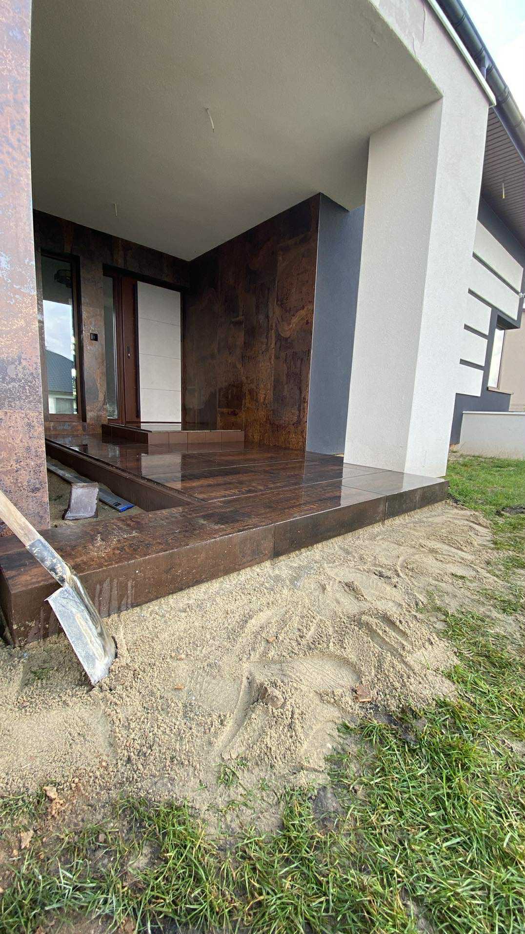 Wykończony taras z brązowych płytek przy wejściu do domu, z widoczną łopatą i piaskiem, sugerujący prace wykończeniowe w toku. Nowoczesny design.