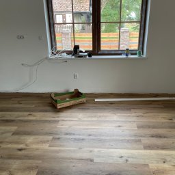 STEEL-DESIGN CONSTRUCTION & INVESTMENT SPÓŁKA Z OGRANICZONĄ ODPOWIEDZIALNOŚCIĄ - Ułożone panele podłogowe w jasnym wnętrzu z oknem. Widoczne listwy przypodłogowe i karton po panelach. Świeżo pomalowane ściany.