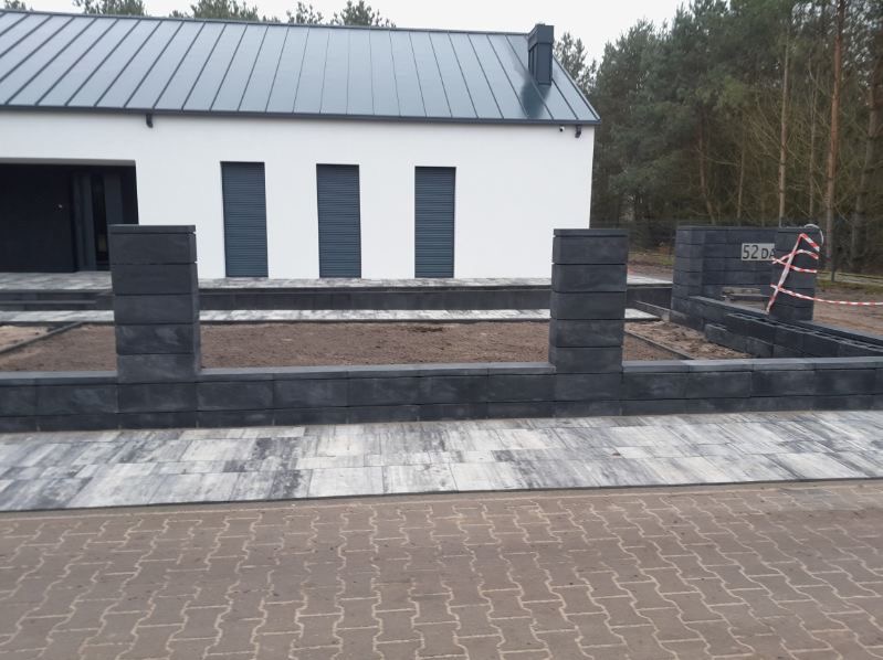 Nowoczesny dom z szarą elewacją i grafitowym dachem, otoczony niskim, modułowym ogrodzeniem z szarej kostki. Ułożona kostka brukowa na podjeździe i ścieżce.