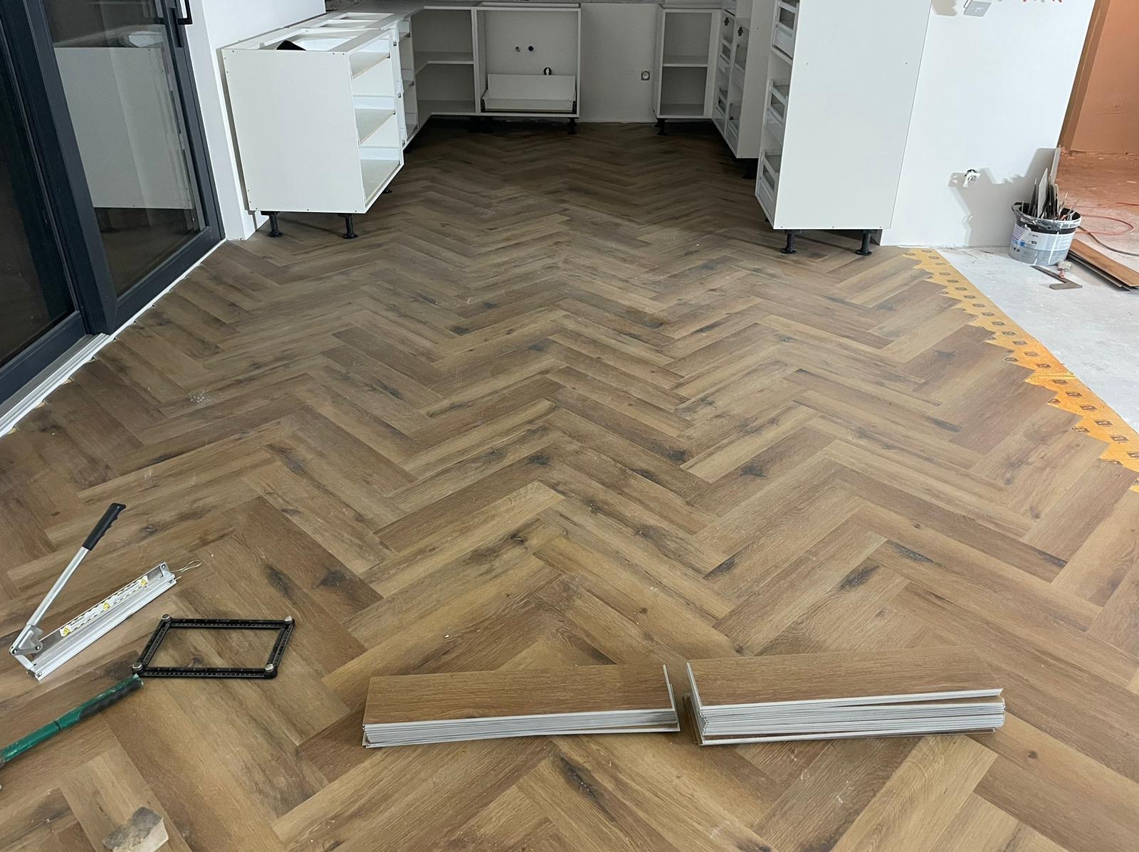 Ułożona podłoga z paneli w jodełkę, widoczne narzędzia i resztki paneli. W tle białe szafki kuchenne bez frontów. Jasne wnętrze w trakcie wykańczania.