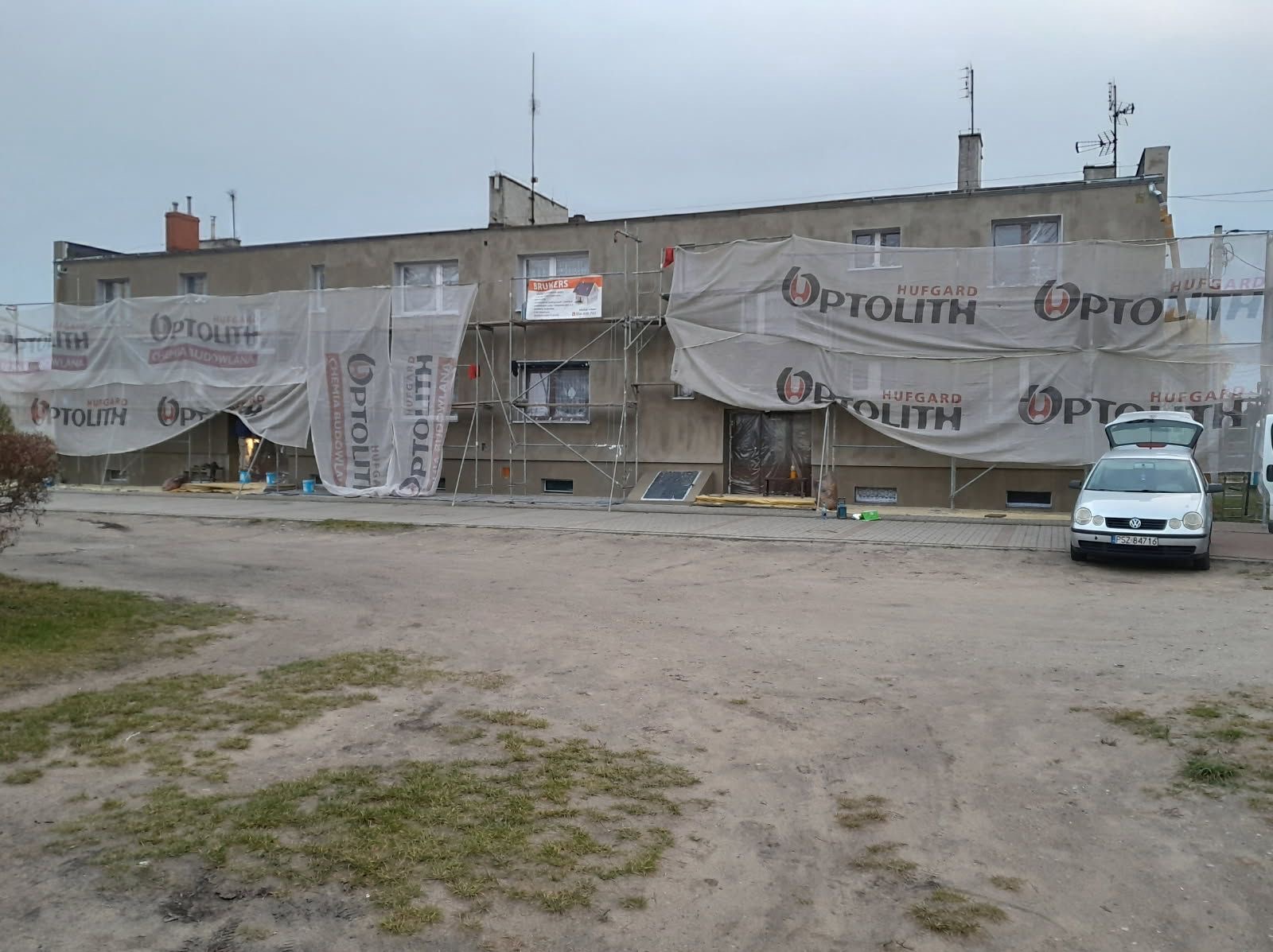 Budynek w trakcie remontu elewacji, osłonięty siatką z logo firmy. Widoczne rusztowania i zaparkowany samochód osobowy. Ujęcie z poziomu gruntu, pochmurny dzień.