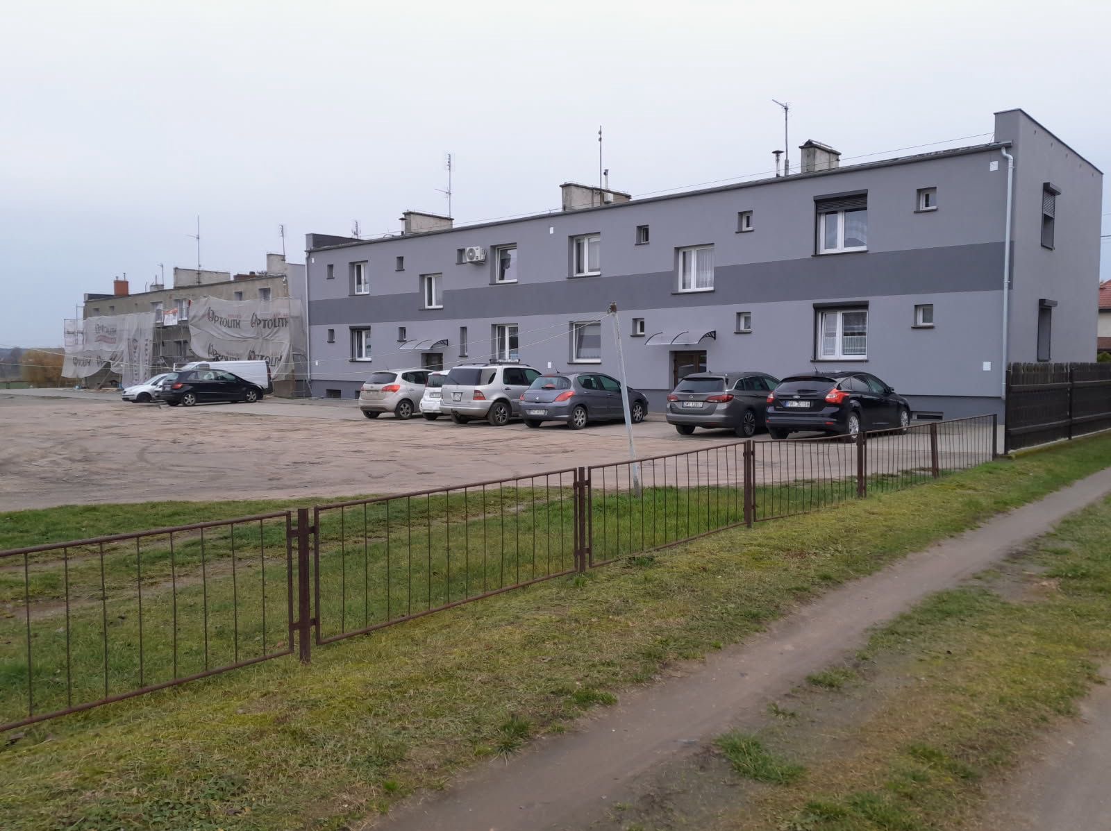 Szary, dwukondygnacyjny budynek po renowacji z ciemniejszym pasem na elewacji, zaparkowane samochody, ogrodzenie i trawnik. Część budynku w rusztowaniach.