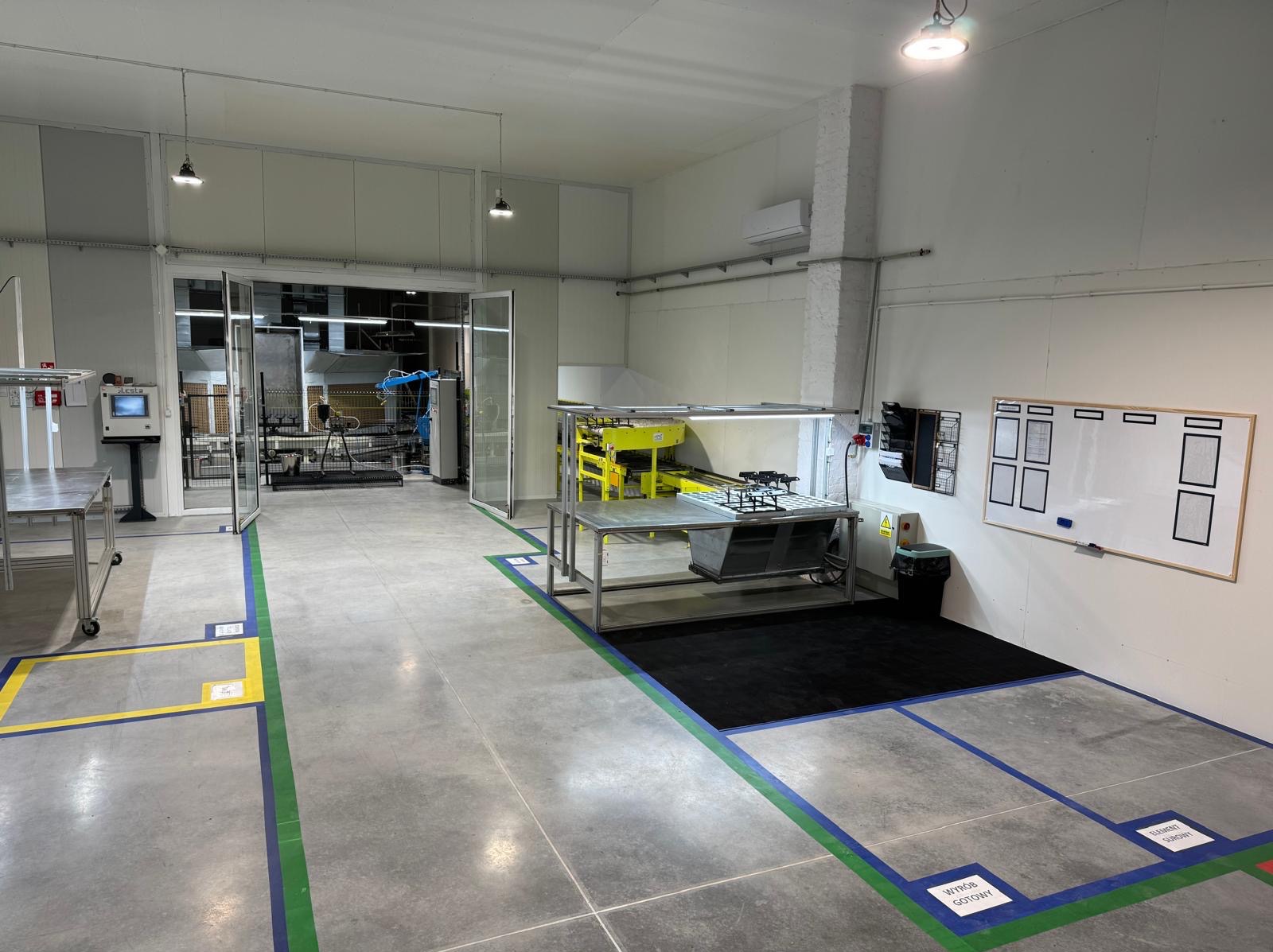 Wnętrze hali produkcyjnej z metalowymi stołami, robotem przemysłowym w tle i wyznaczonymi strefami na podłodze. Biała tablica suchościeralna na ścianie.