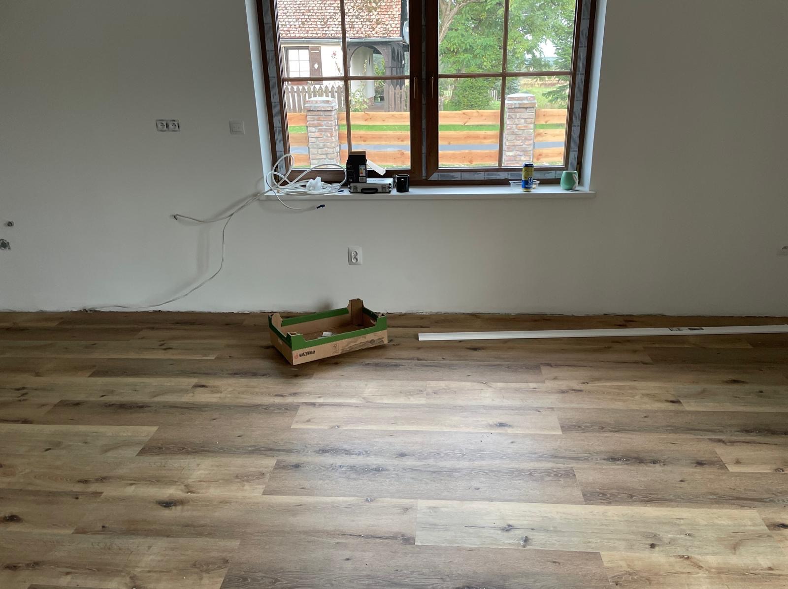Ułożone panele podłogowe w jasnym wnętrzu z oknem. Widoczne listwy przypodłogowe i karton po panelach. Świeżo pomalowane ściany.