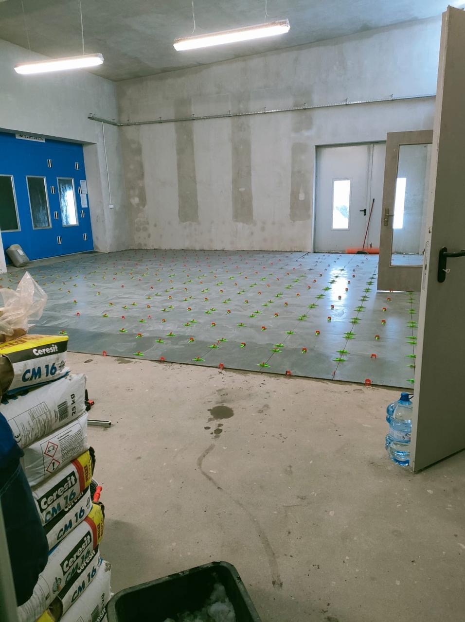 Układanie płytek podłogowych w dużym pomieszczeniu z użyciem systemu poziomowania. Widoczne worki z klejem Cerisit CM 16 i butelki z wodą.