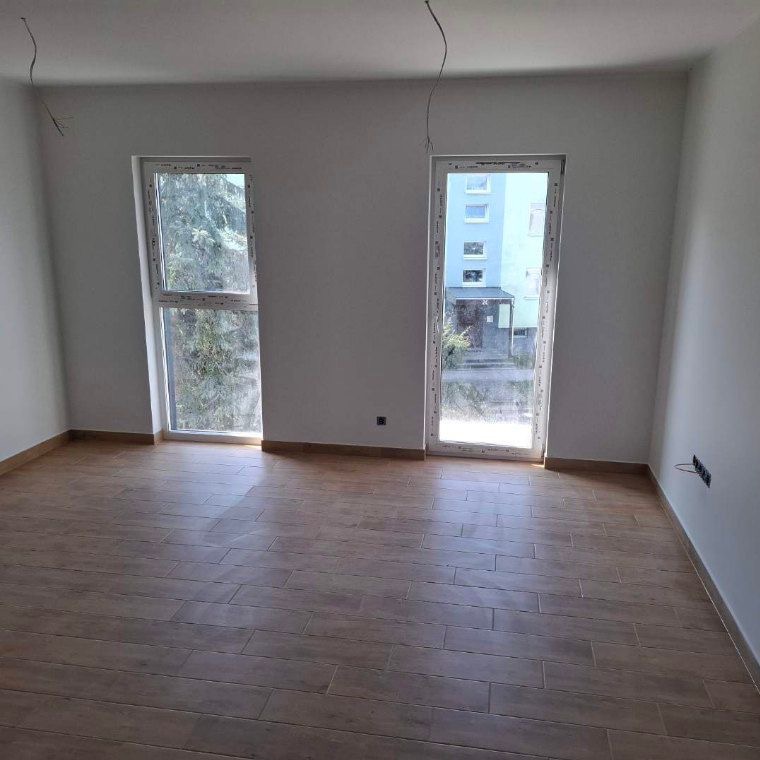 Jasne wnętrze z drewnopodobną podłogą z płytek, dwa okna z widokiem na zewnątrz, białe ściany i wystające kable. Minimalistyczny design.