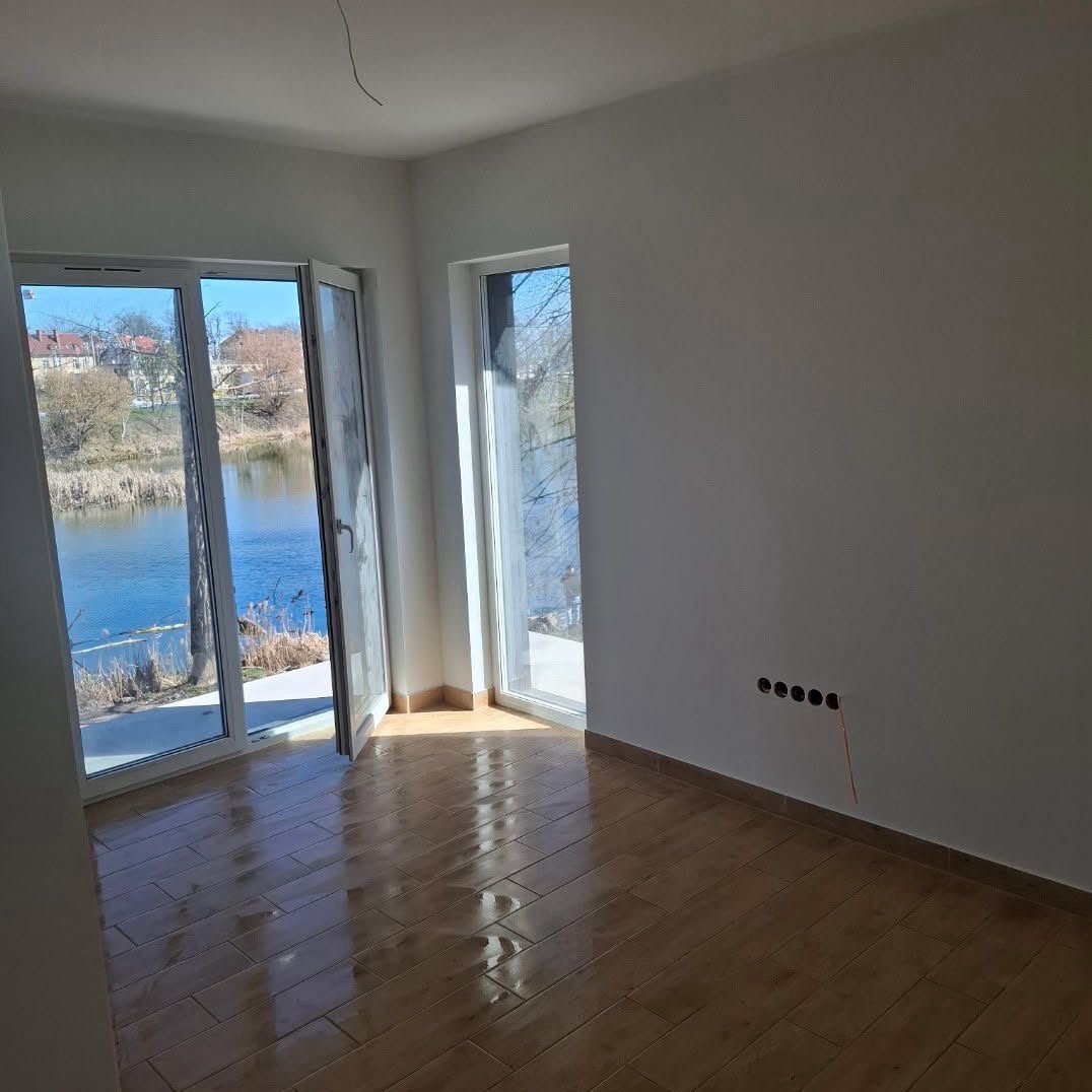 Jasne wnętrze po remoncie: parkiet w odcieniach brązu, białe ściany z gniazdkami, okno z widokiem na jezioro i otwarty balkon. Minimalistyczny design.