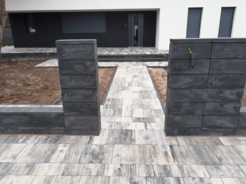 Nowoczesne wejście do domu z szarej kostki brukowej. Kolumny z ciemnych bloczków betonowych. Minimalistyczny design, surowe wykończenie.