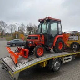 Pomarańczowy traktor Kubota ST-30 z pługiem na ż&oacute;łtej lawecie pomocy drogowej Strumyk, widok z boku na szarym tle parkingu i drzew bez liści.