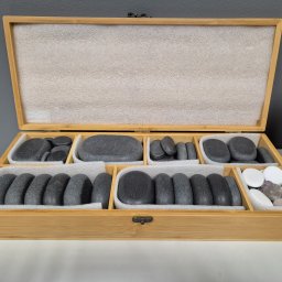 Sana Persona Piotr Ścigała - Otwarta drewniana skrzynka z przegródkami, w których znajdują się kamienie do masażu w różnych kształtach i kolorach, ułożone na białym podłożu.