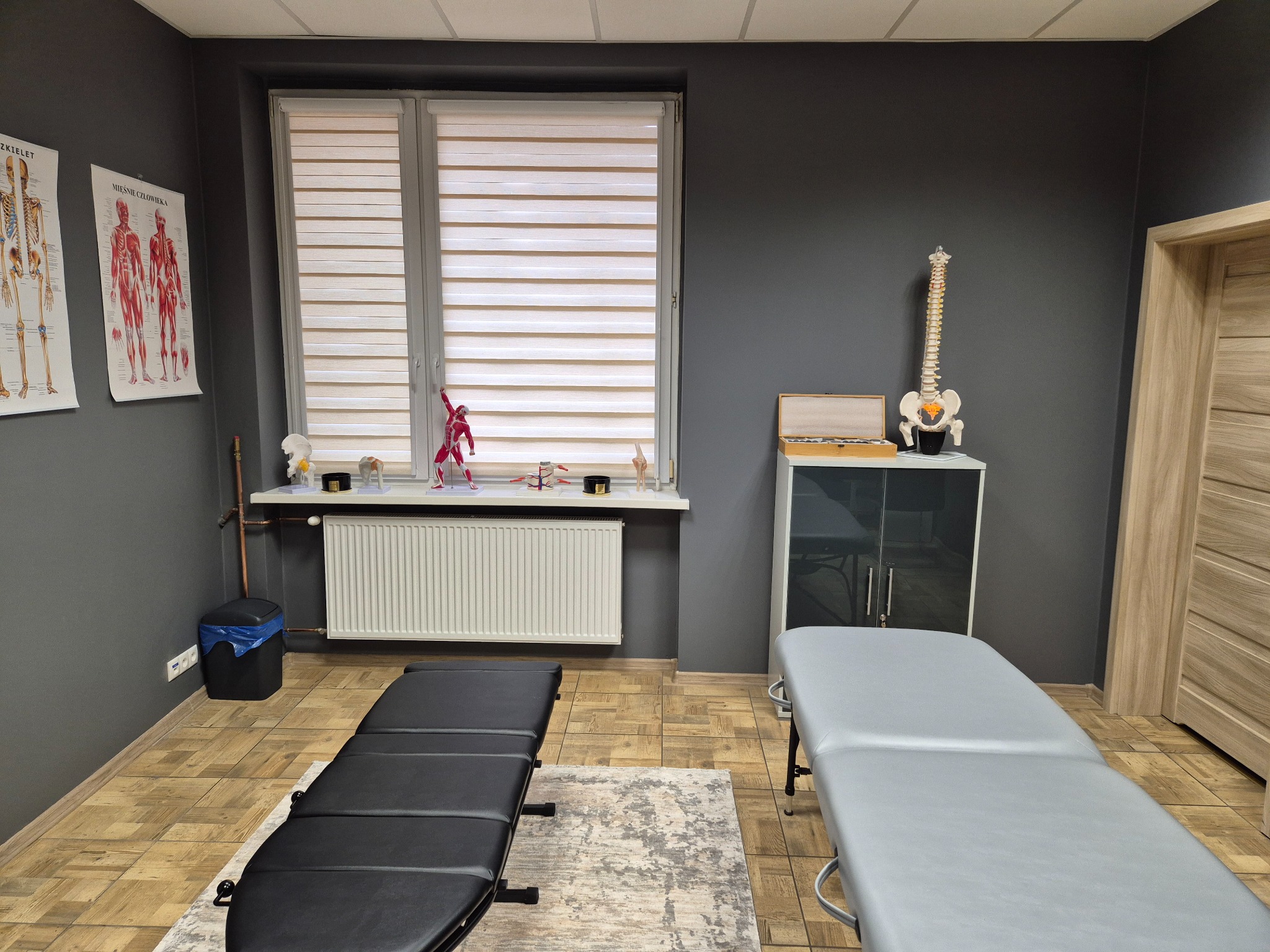 Gabinet z dwoma leżankami do masażu, model szkieletu na szafce, plakaty anatomiczne na ścianie i drewniana podłoga.