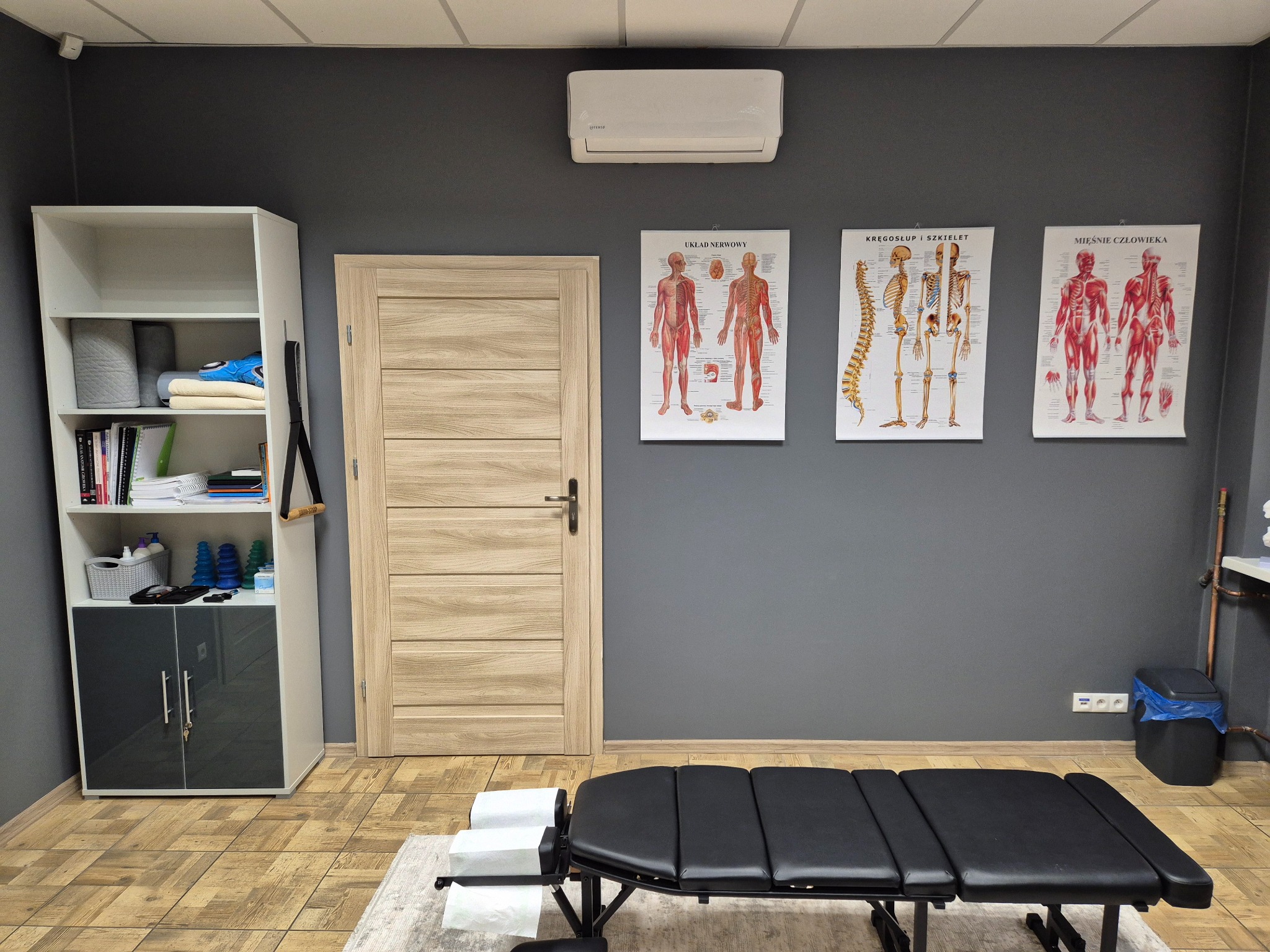 Gabinet z czarnym stołem do masażu, plakatami anatomicznymi na szarej ścianie, szafką i klimatyzatorem. Jasne drewno drzwi i podłogi. Widok na wnętrze.