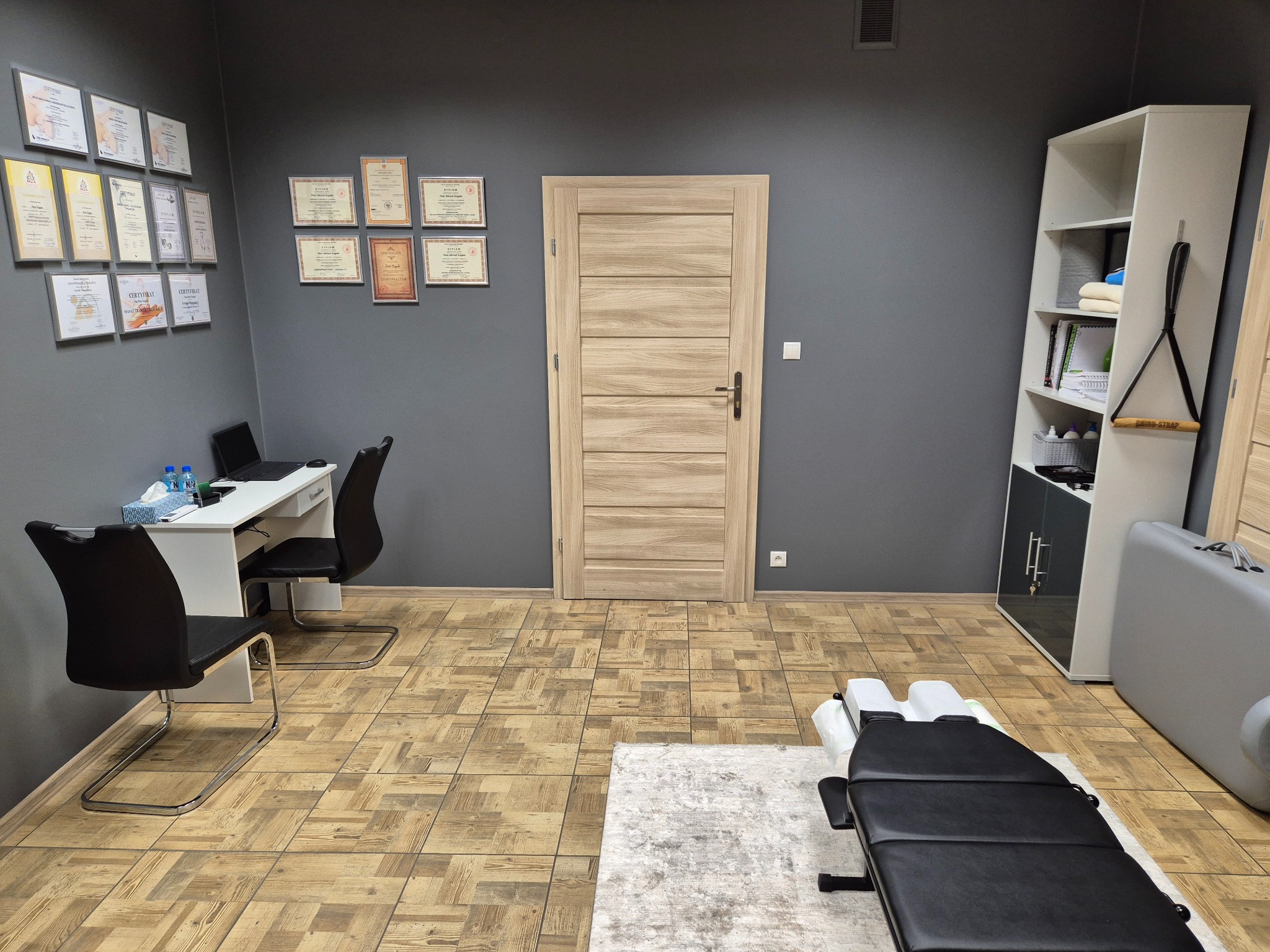 Gabinet z biurkiem, dyplomami na ścianie i czarnym stołem do zabiegów na dywanie. W tle regał i drzwi. Stonowana kolorystyka wnętrza.