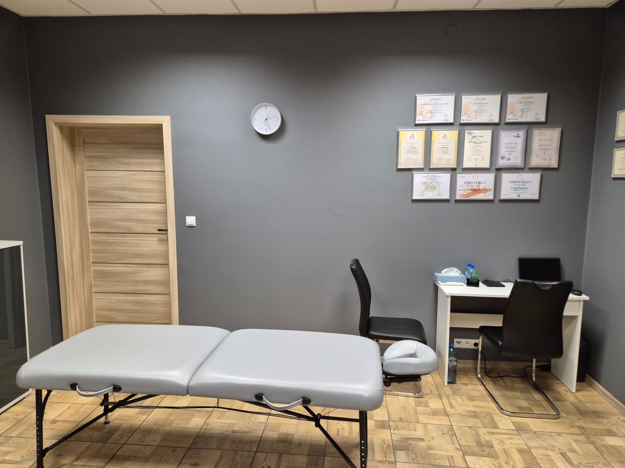 Jasne wnętrze gabinetu masażu z szarymi ścianami, zegarem, dyplomami, biurkiem i szarym stołem do masażu z regulacją wysokości.
