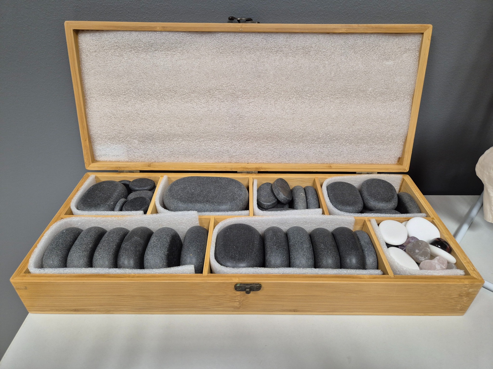 Otwarta drewniana skrzynka z przegródkami, w których znajdują się kamienie do masażu w różnych kształtach i kolorach, ułożone na białym podłożu.