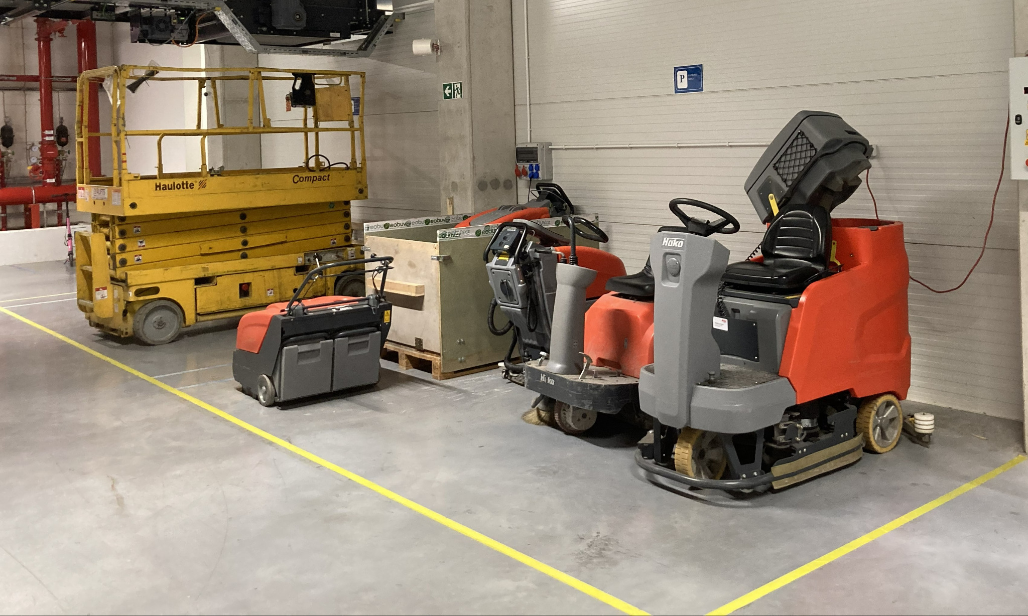 Różne maszyny czyszczące i podnośnik koszowy w hali z betonową podłogą i żółtymi liniami. Widoczne urządzenia Hako i Haulotte.