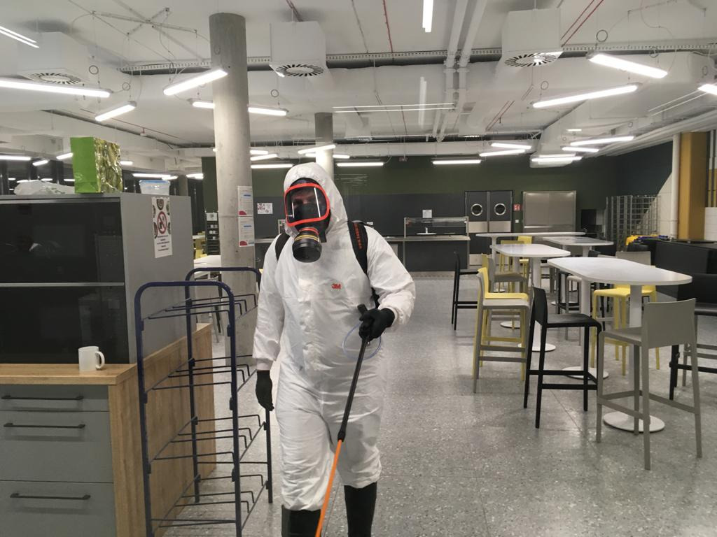 Dezynfekcja biura: osoba w kombinezonie ochronnym i masce rozpyla środek dezynfekujący w przestrzeni biurowej z meblami i stołami. Widoczne oświetlenie sufitowe.