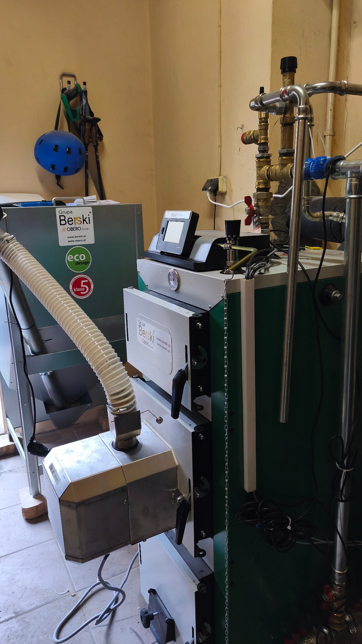 Nowoczesny piec na pellet marki Berski z podajnikiem, widoczne rury instalacji CO, niebieski kask i kijki narciarskie na ścianie w tle.