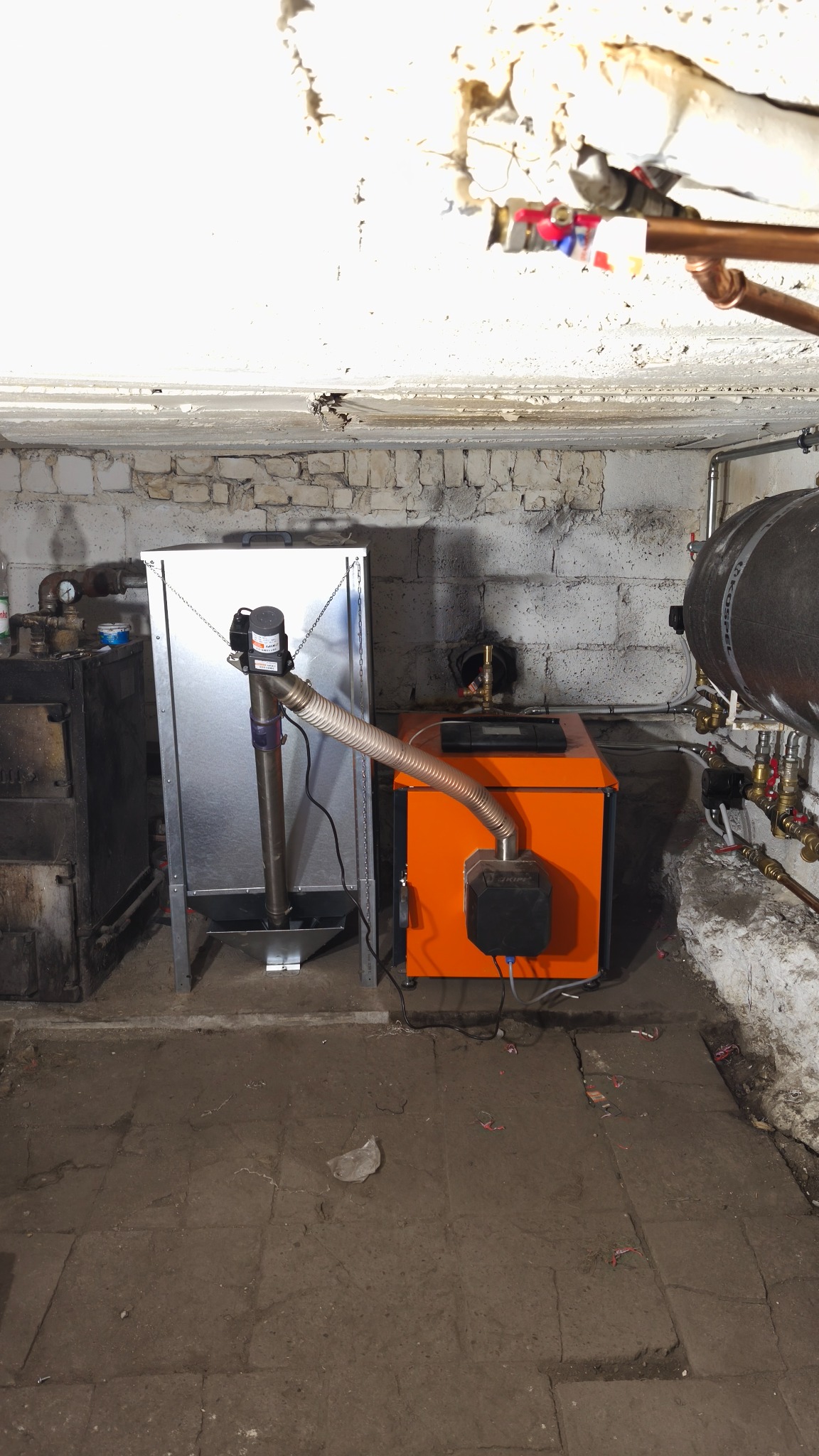 Pomarańczowy piec CO z podajnikiem w kotłowni, widoczne rury i instalacje. Surowe ściany z cegły i betonowa podłoga. Oświetlenie górne.