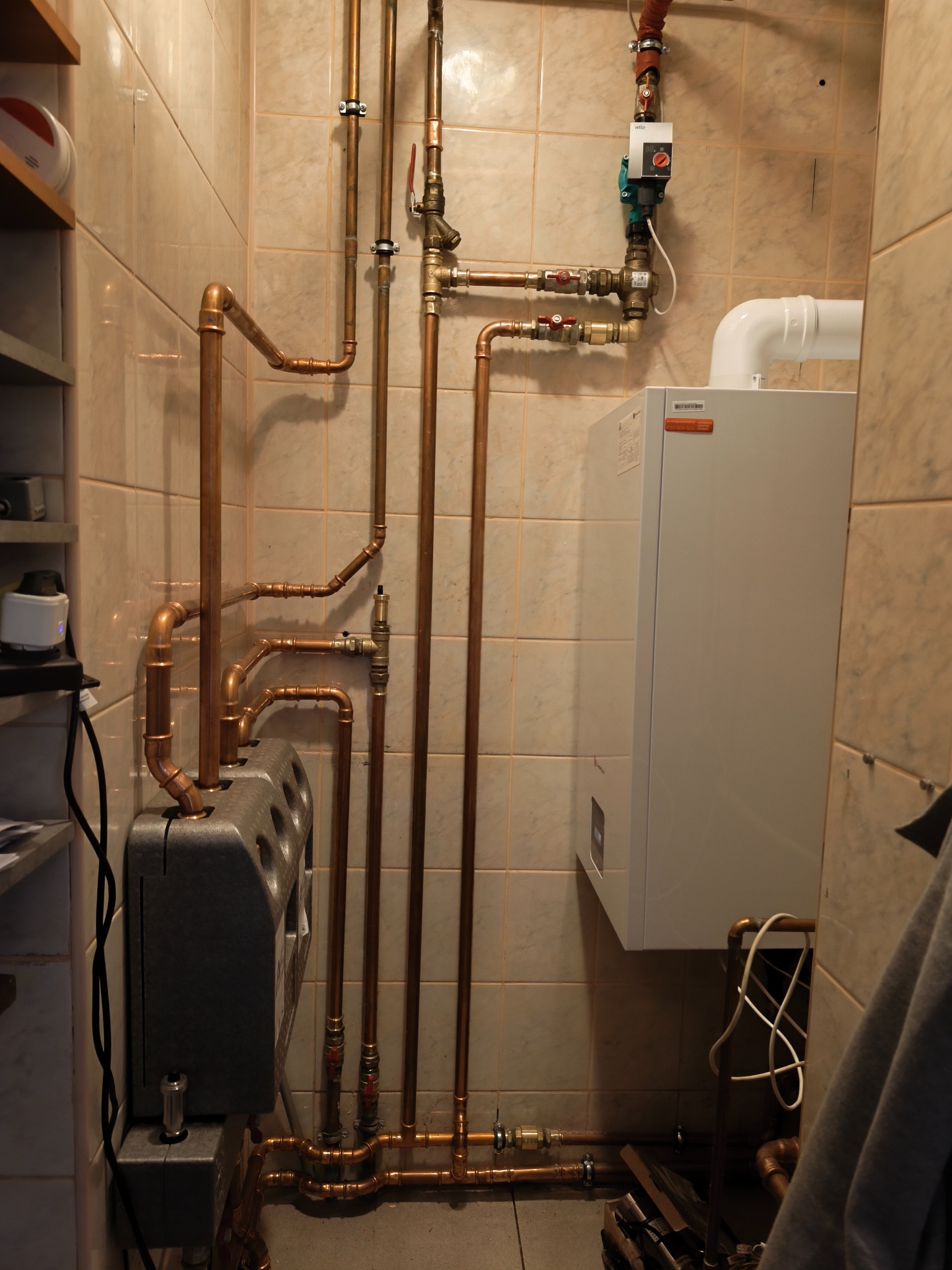 Instalacja CO z piecem gazowym kondensacyjnym, rurami miedzianymi i zaworami, zamontowana na kafelkowanej ścianie w pomieszczeniu technicznym. Widoczne elementy sterujące.