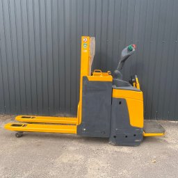 PRO LIFT Miłosz Brodzik - Żółto-szary wózek paletowy elektryczny Jungheinrich EJE 116, widok z boku na tle szarej ściany z blachy falistej i fragmentu ogrodzenia z siatki.