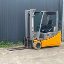 PRO LIFT Miłosz Brodzik - Żółto-szary wózek widłowy Jungheinrich EFG 220 stoi na zewnątrz, z widocznymi widłami i oponami. Tło stanowi szara ściana i siatka ogrodzeniowa.