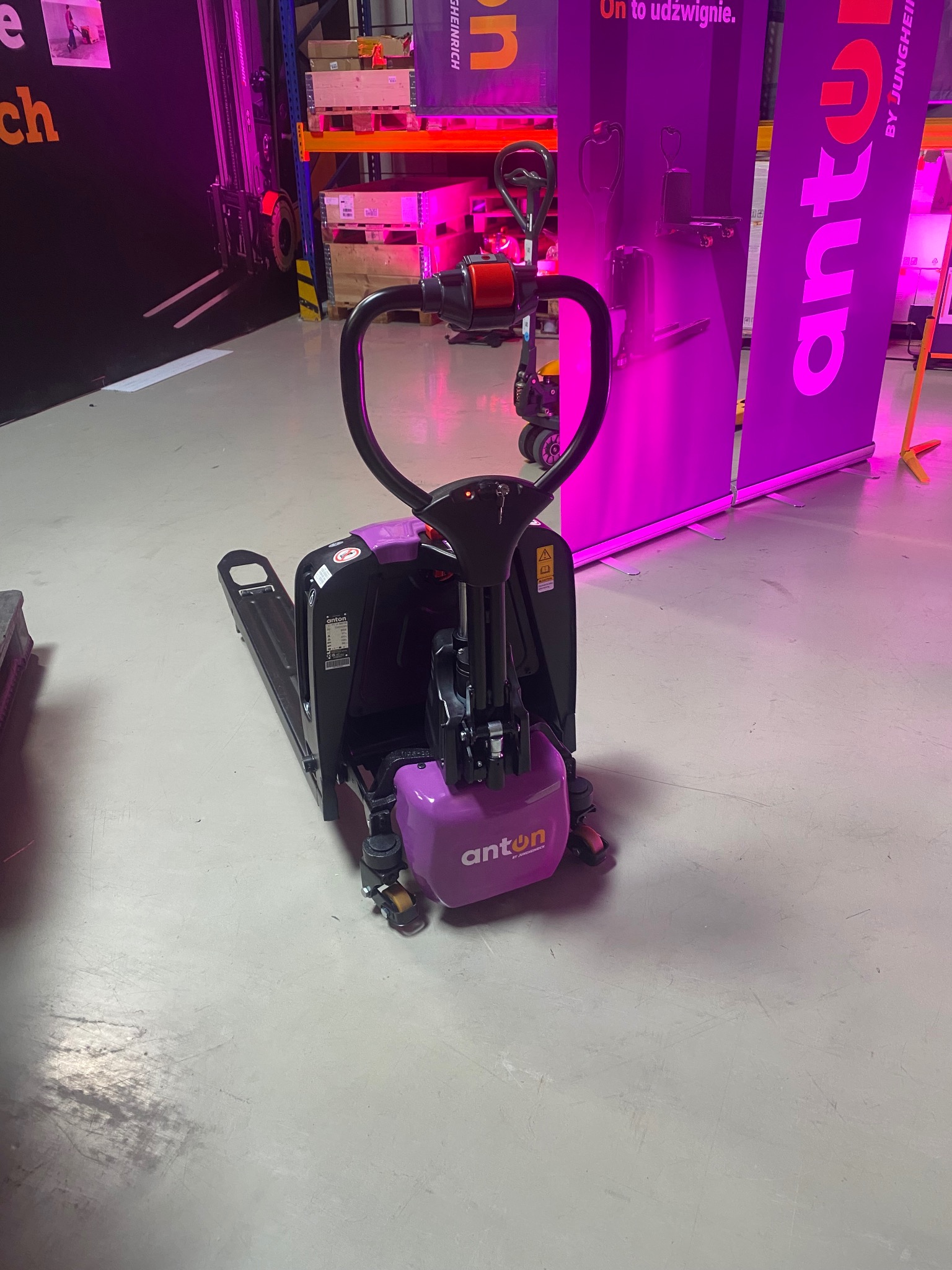 Elektryczny wózek paletowy Anton by Jungheinrich w fioletowej kolorystyce, prezentowany na tle magazynu z regałami i banerami reklamowymi.