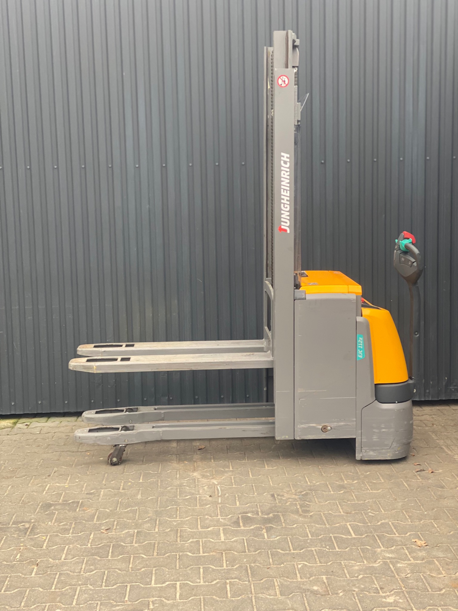 Elektryczny wózek paletowy Jungheinrich EJC 112z, widok z boku na zewnątrz, szaro-żółta kolorystyka, na tle szarej, blaszanej ściany.
