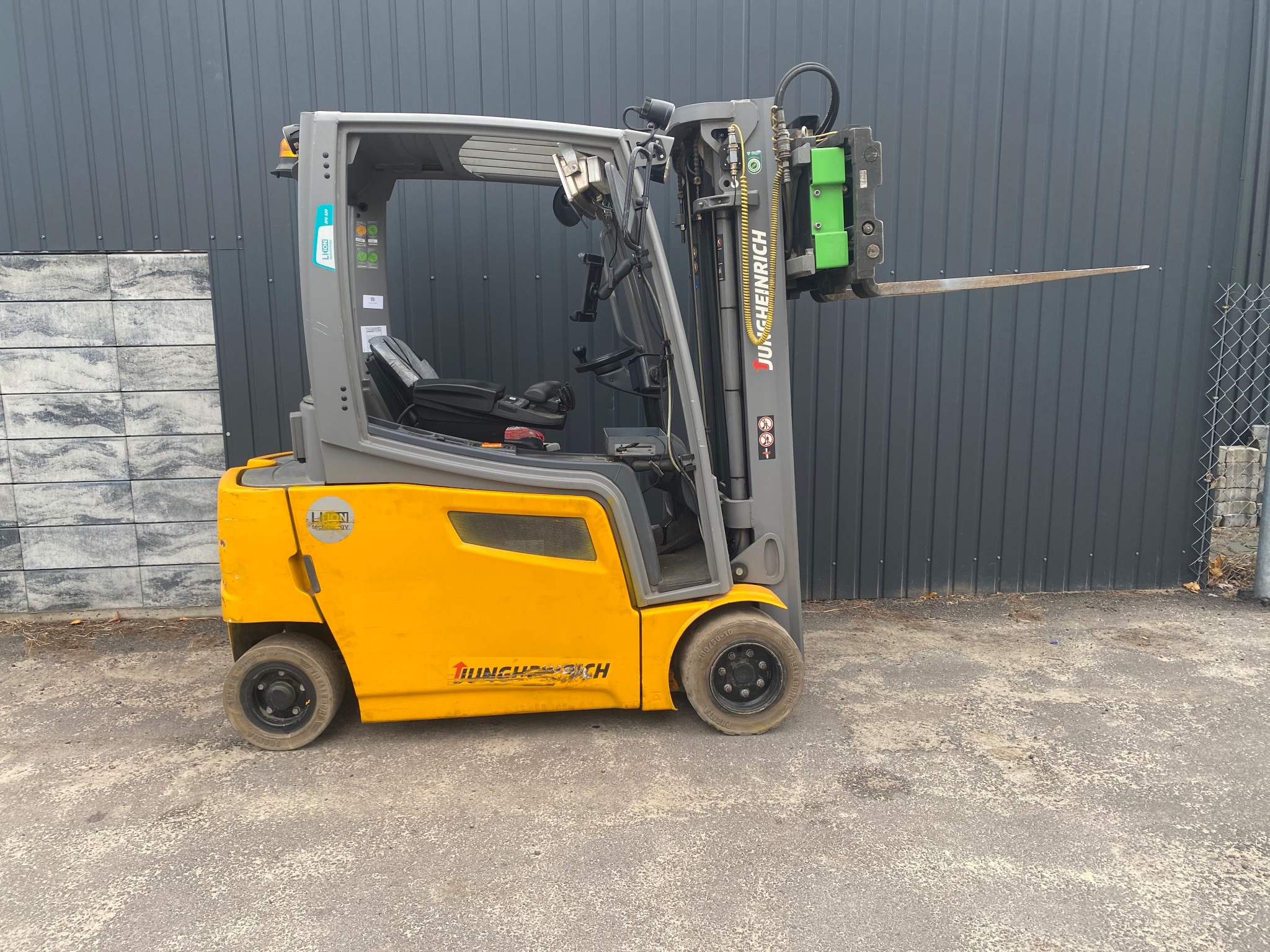 Żółto-szary wózek widłowy Jungheinrich na zewnątrz, z widocznymi detalami konstrukcyjnymi i logo. Tło stanowi szara ściana i fragment chodnika.