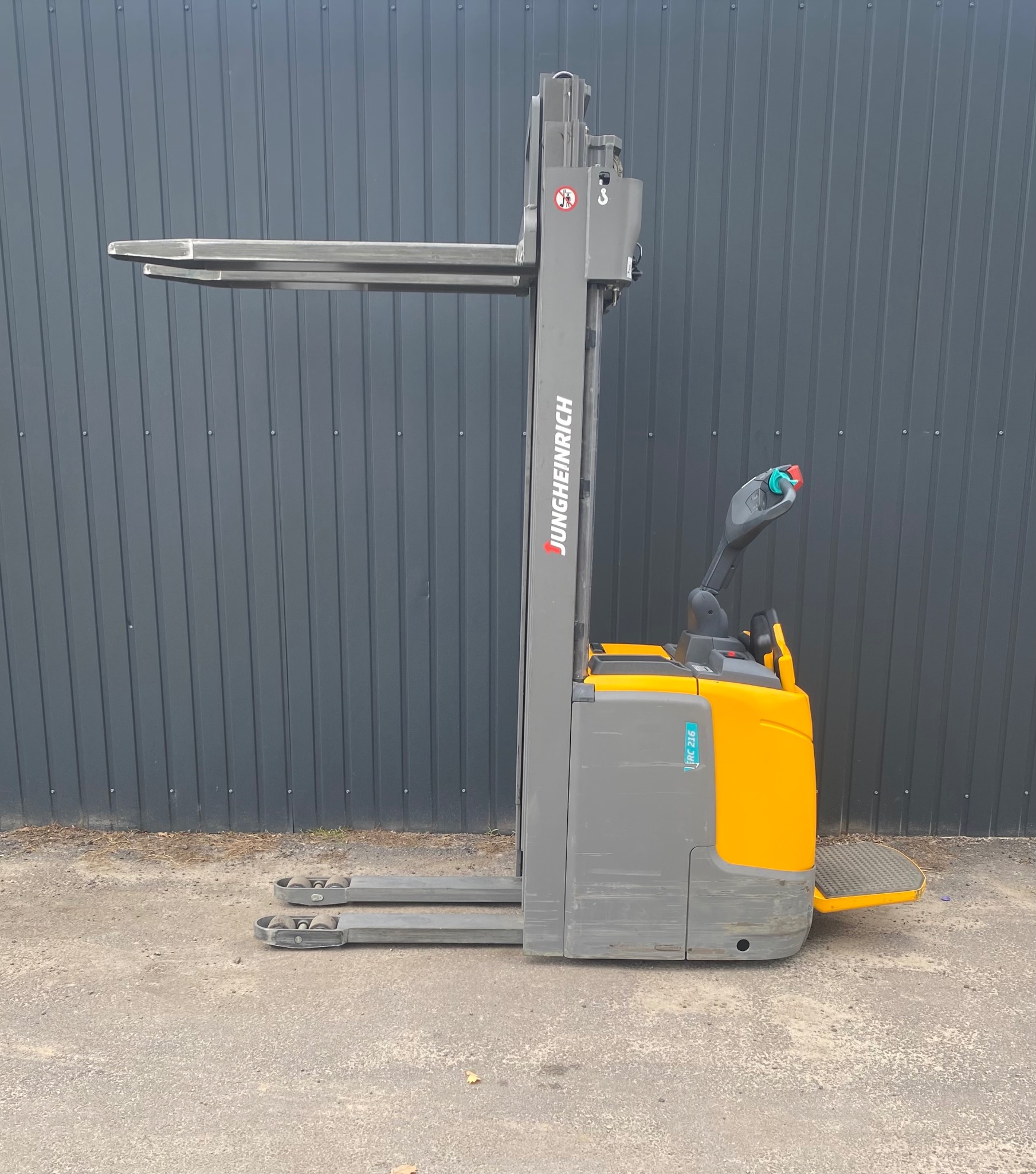 Elektryczny wózek widłowy Jungheinrich z wysuwanym masztem, widok z boku na tle szarej, blaszanej ściany. Widoczne detale konstrukcyjne i oznaczenia modelu.