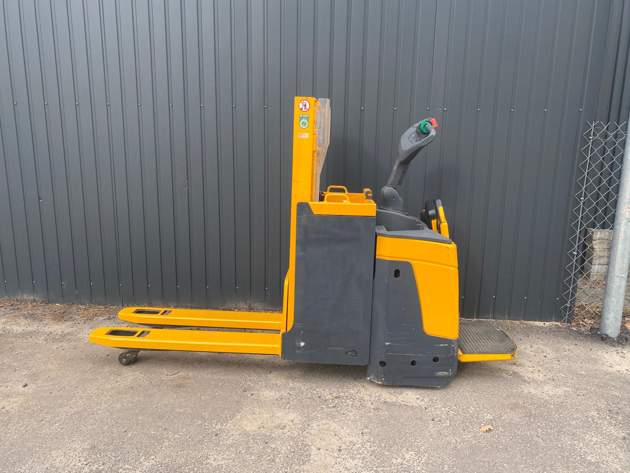 Żółto-szary wózek paletowy elektryczny Jungheinrich EJE 116, widok z boku na tle szarej ściany z blachy falistej i fragmentu ogrodzenia z siatki.
