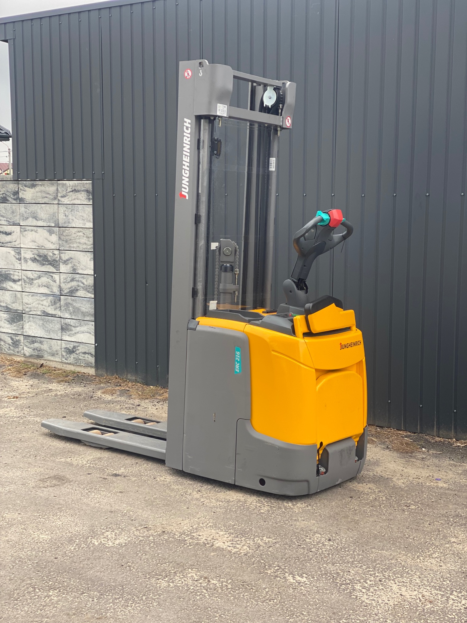 Żółto-szary wózek widłowy Jungheinrich ERC 216 na zewnątrz, na tle szarej ściany z blachy trapezowej i fragmentu muru z szarej kostki. Widok z boku, lekko z dołu.