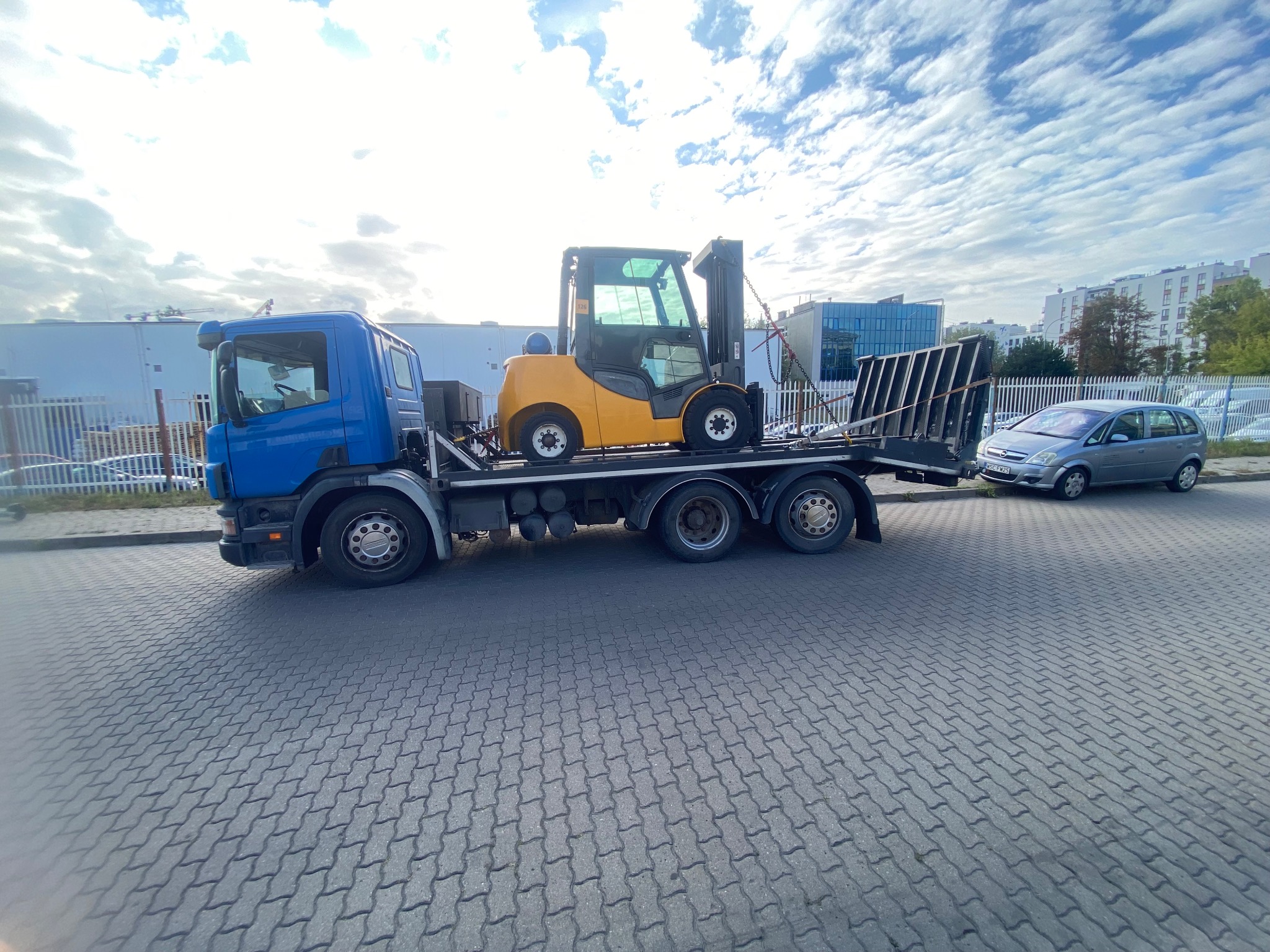 Żółty wózek widłowy na lawecie niebieskiej ciężarówki, transportowany na tle miejskiego krajobrazu z budynkami i pochmurnym niebem. Widoczny szary samochód osobowy.