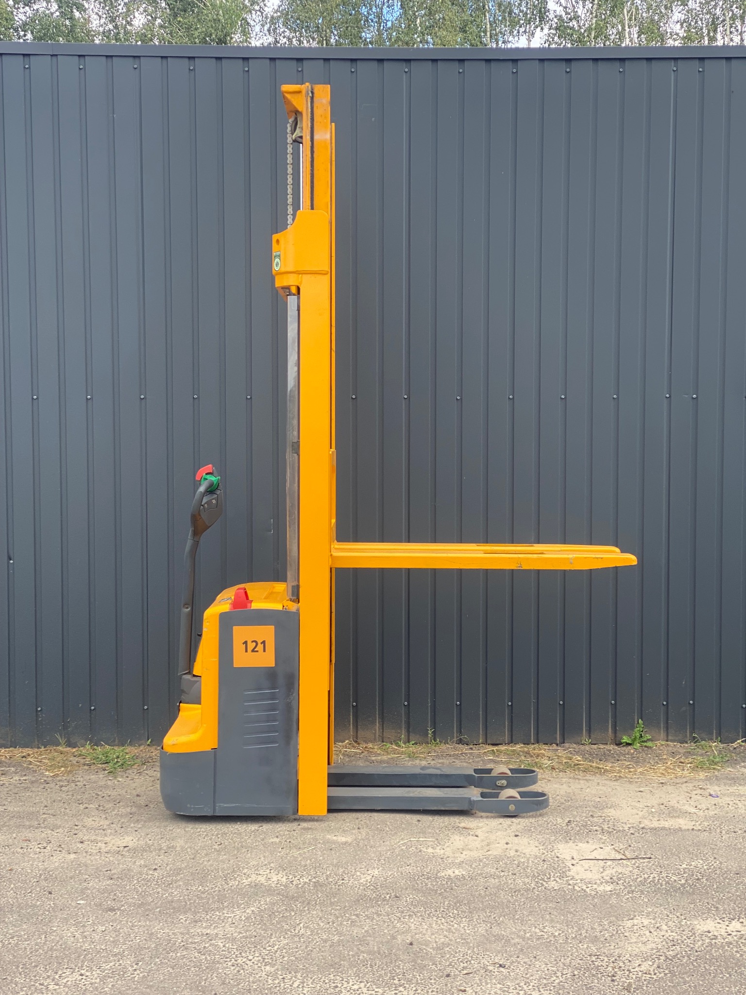 Żółto-szary wózek paletowy elektryczny z numerem 121 na tle szarej, blaszanej ściany i drzew. Widok z boku, na zewnątrz.