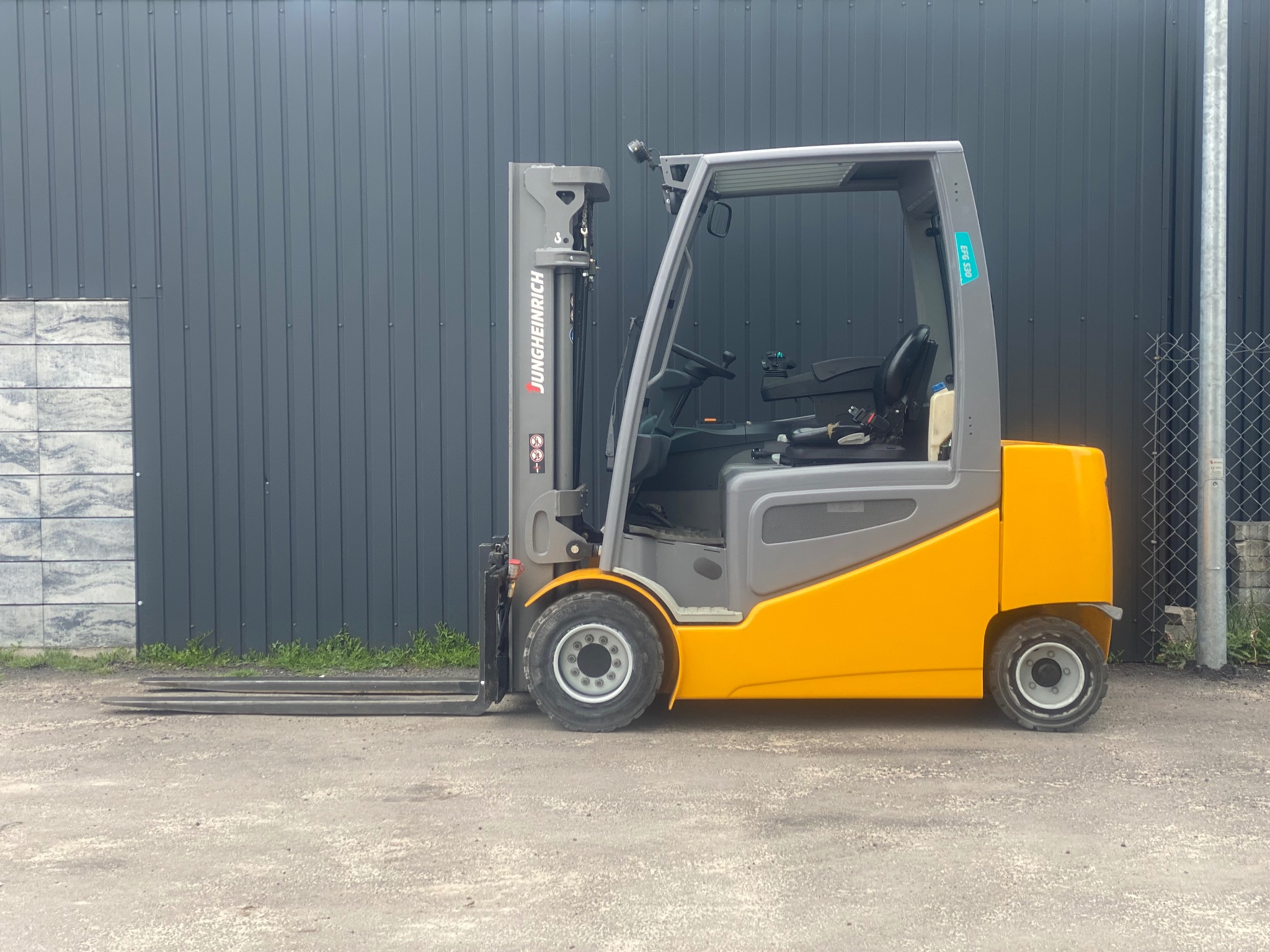 Żółto-szary wózek widłowy Jungheinrich EFG 530 na zewnątrz, na tle szarej ściany z blachy trapezowej. Widoczne detale konstrukcyjne i opony wózka.