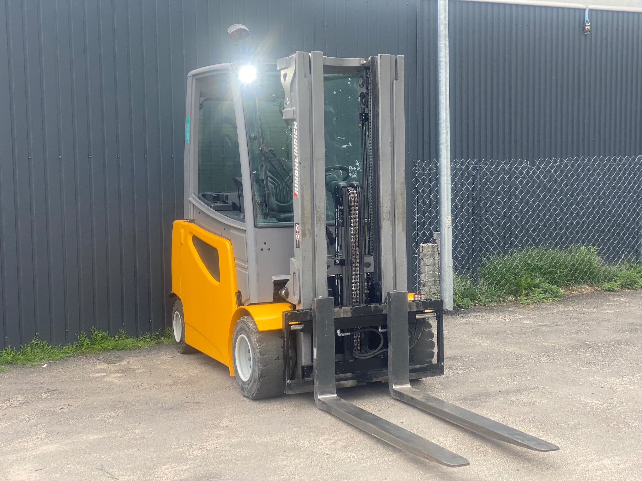 Żółto-szary wózek widłowy elektryczny Jungheinrich, stojący na zewnątrz na betonowym podłożu, z widocznymi widłami i oświetleniem.