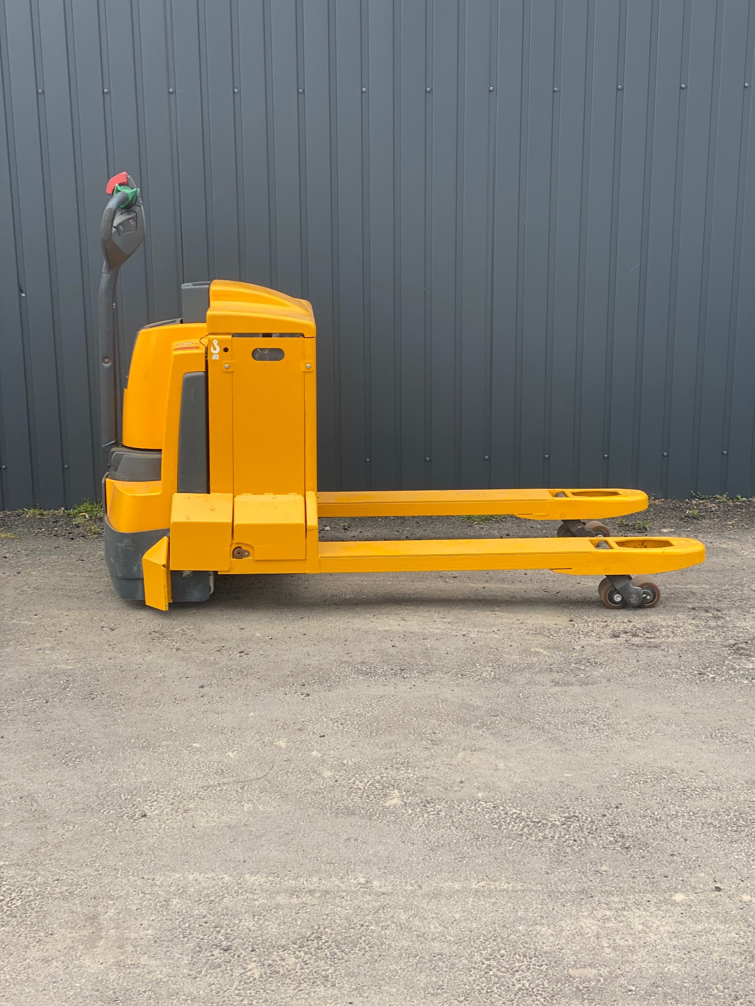 Żółty, elektryczny wózek paletowy Jungheinrich EJE 116, widok z boku na szarym tle. Wózek stoi na betonowej powierzchni.