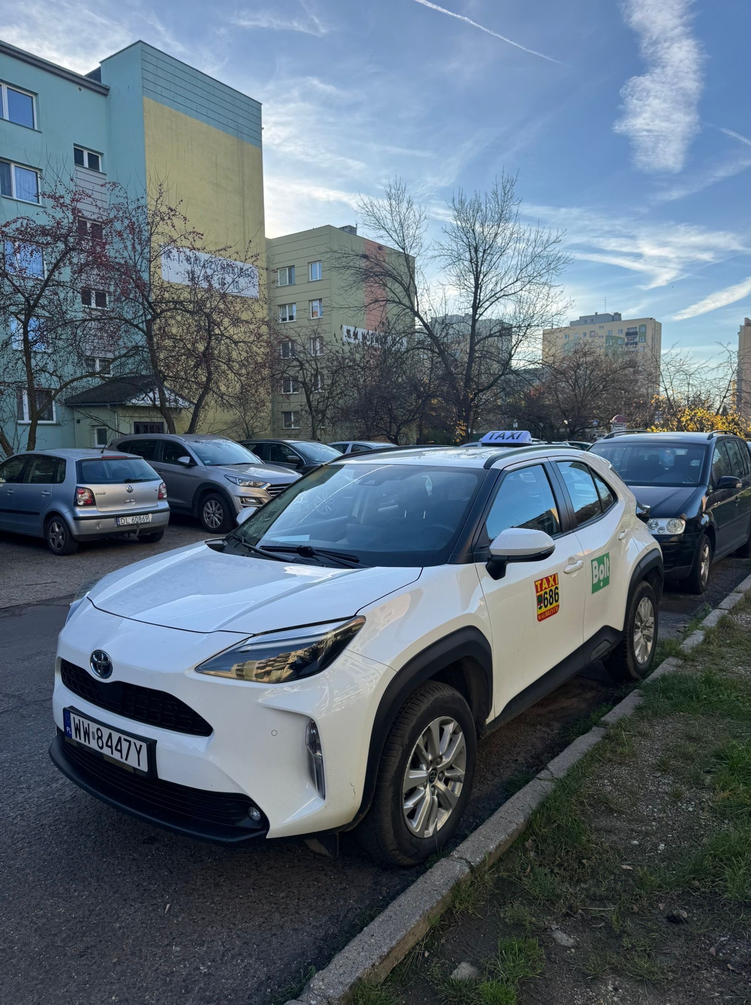 Biały samochód Toyota Yaris z oznaczeniami Taxi Bolt zaparkowany na ulicy, na tle bloku mieszkalnego i innych zaparkowanych aut. Tablica rejestracyjna widoczna.