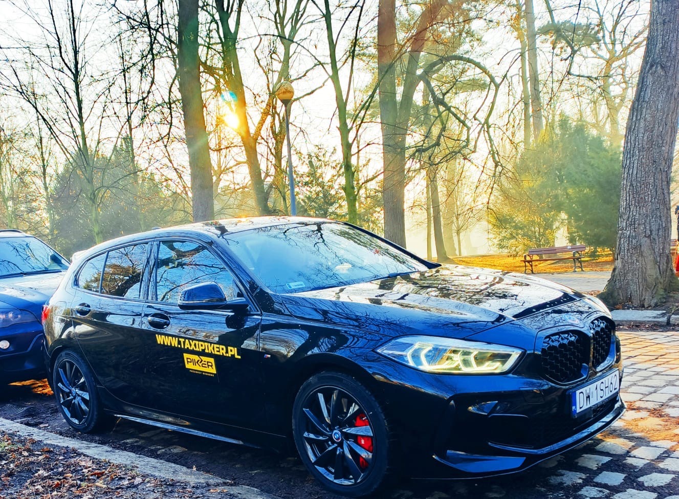 Czarny hatchback z żółtym logo Taxi Piker na tle parku w słoneczny dzień. Widoczne detale: felgi, czerwone zaciski hamulcowe, tablica rejestracyjna. Częściowo widoczny drugi samochód.