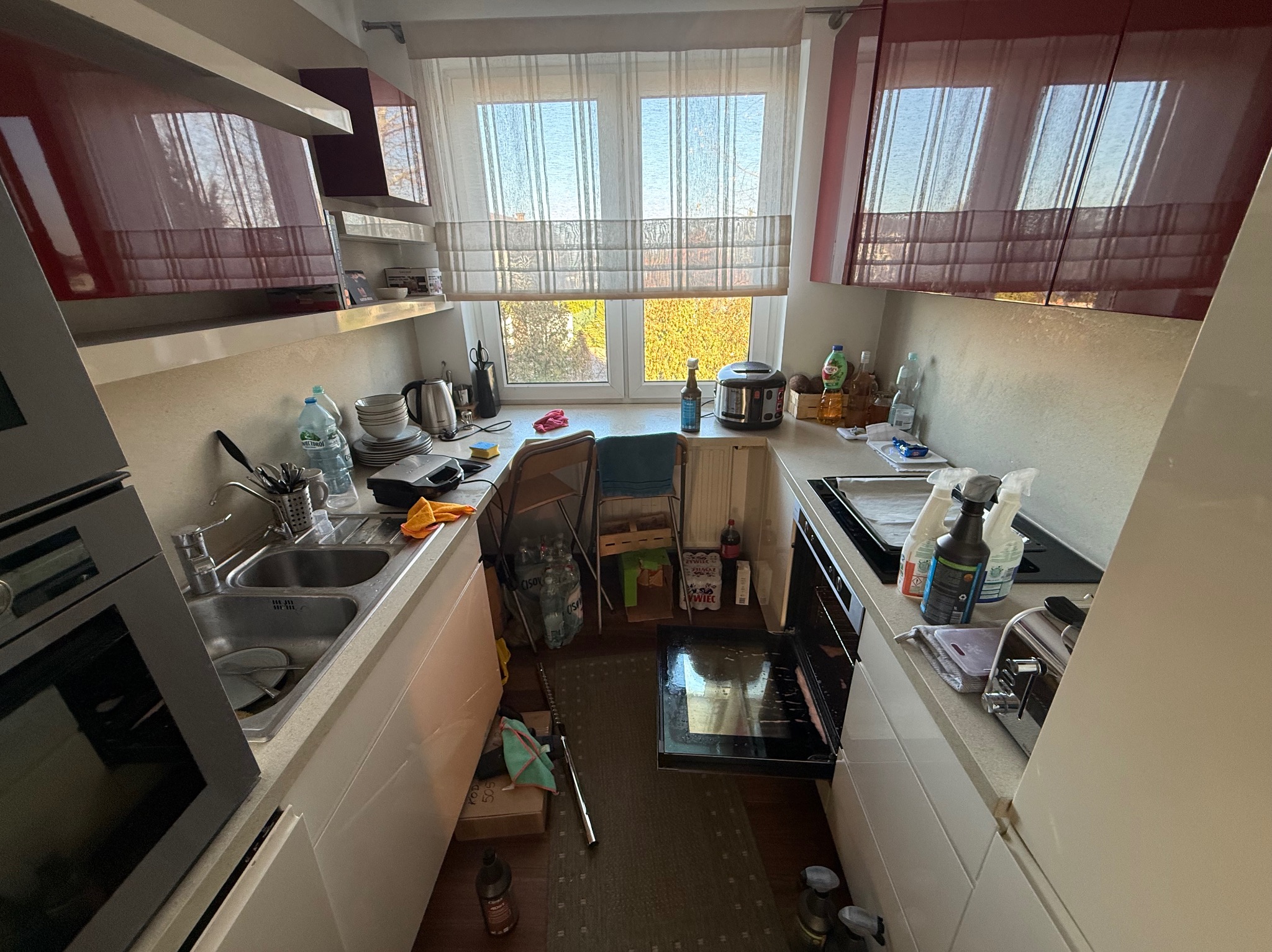 Kuchenny nieład: naczynia w zlewie, butelki i sprzęty na blacie, otwarty piekarnik. Widok na okno zza zagraconej przestrzeni.