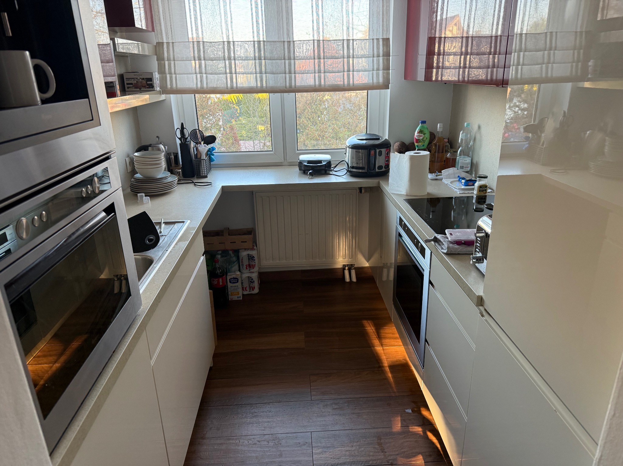 Jasna kuchnia w kształcie litery U z drewnianą podłogą, widok na okno z roletami rzymskimi. Na blacie kuchennym akcesoria i urządzenia. Promienie słoneczne na podłodze.