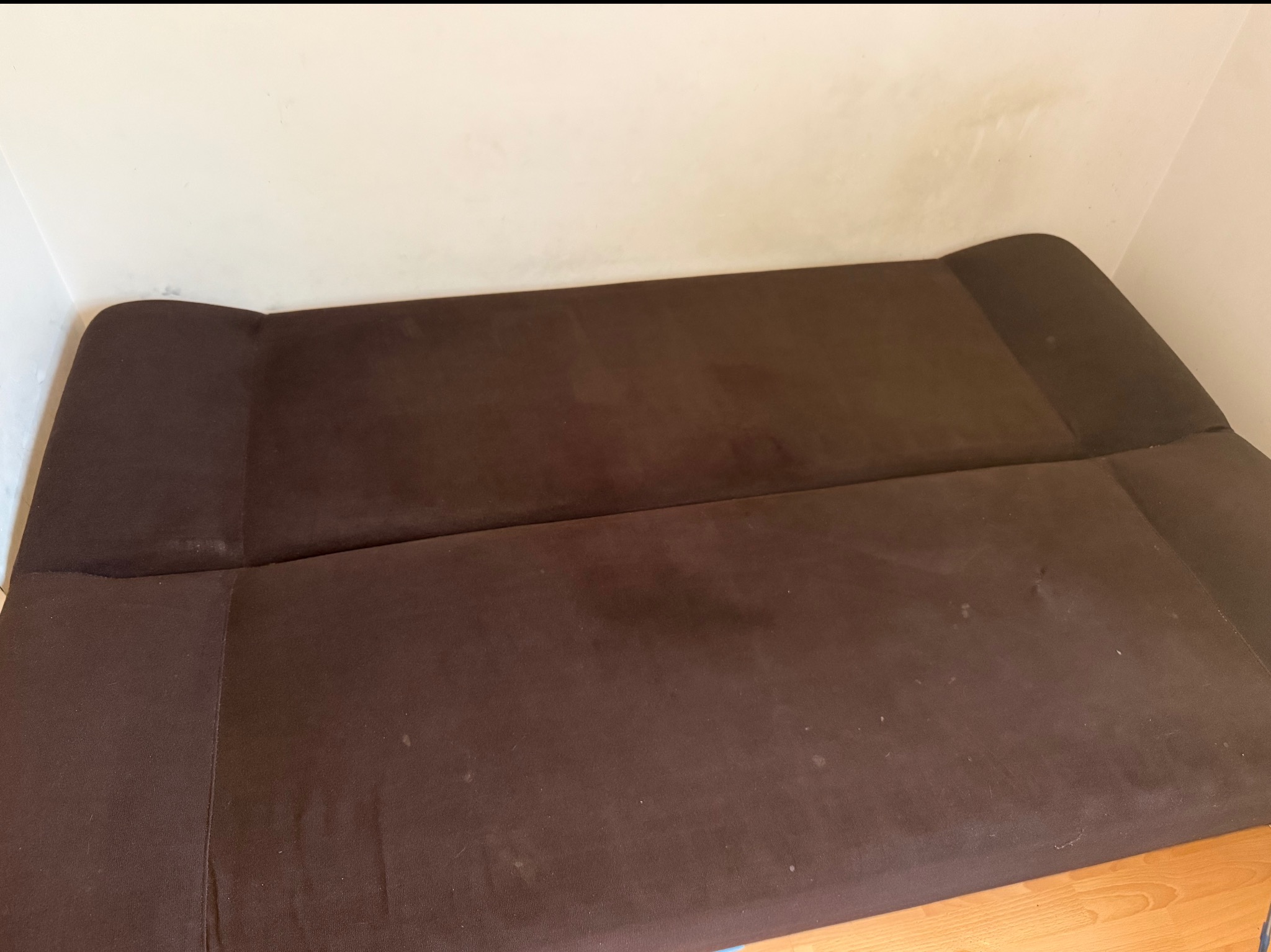 Ciemnobrązowa, rozłożona kanapa z widocznymi plamami na tapicerce, ustawiona pod ścianą. Widoczny fragment podłogi. Ujęcie z góry.