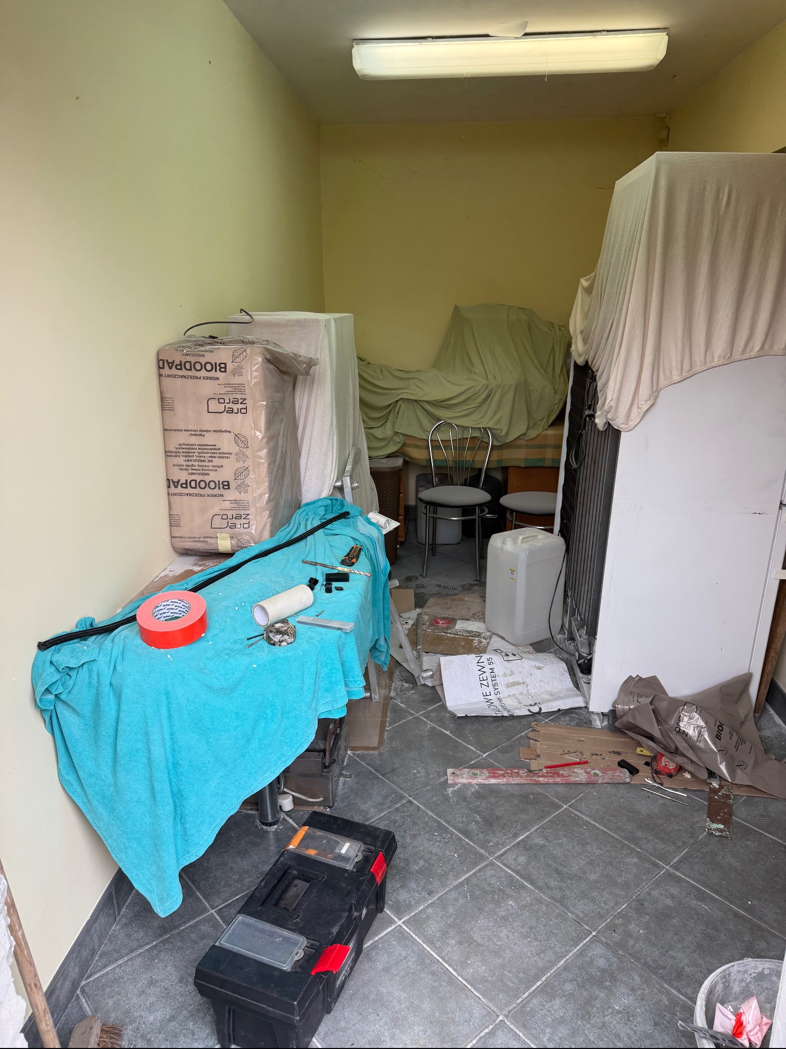 Pomieszczenie zagracone przedmioty pod przykryciem, narzędzia na stole, kartony, wiadro, podłoga w szarą kratę. Widok na wnętrze w trakcie opróżniania.