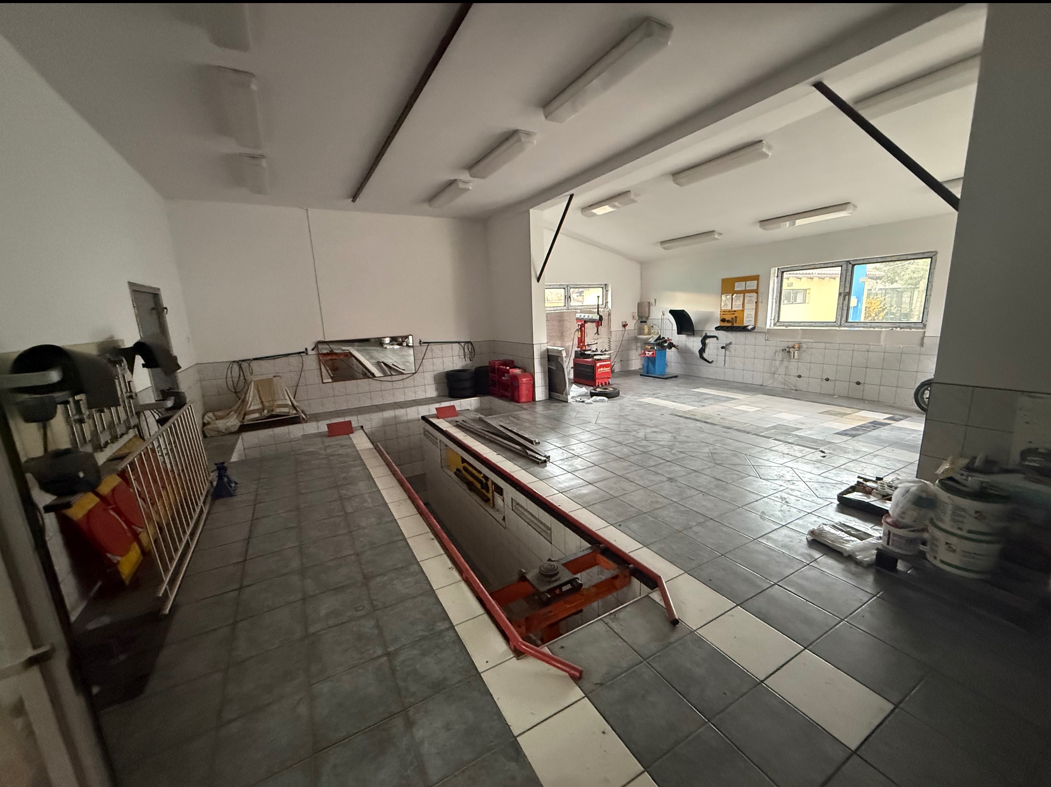 Wnętrze warsztatu samochodowego z kanałem naprawczym, narzędziami i wyposażeniem. Widoczne płytki na ścianach i podłodze, oświetlenie sufitowe. Ujęcie z perspektywy szerokokątnej.