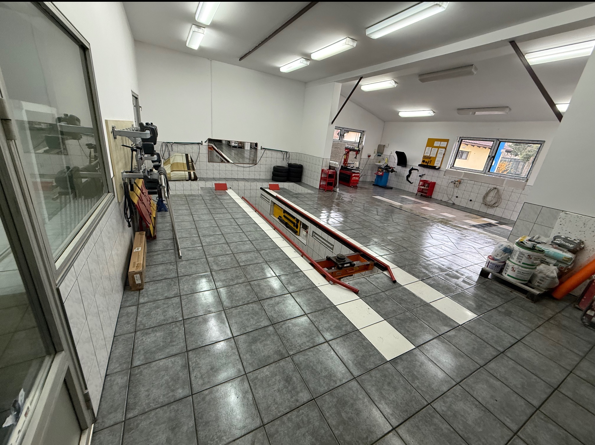 Wnętrze warsztatu samochodowego z podnośnikiem, narzędziami i oponami. Widoczne białe ślady po czyszczeniu na szarej podłodze z płytek. Jasne oświetlenie sufitowe.