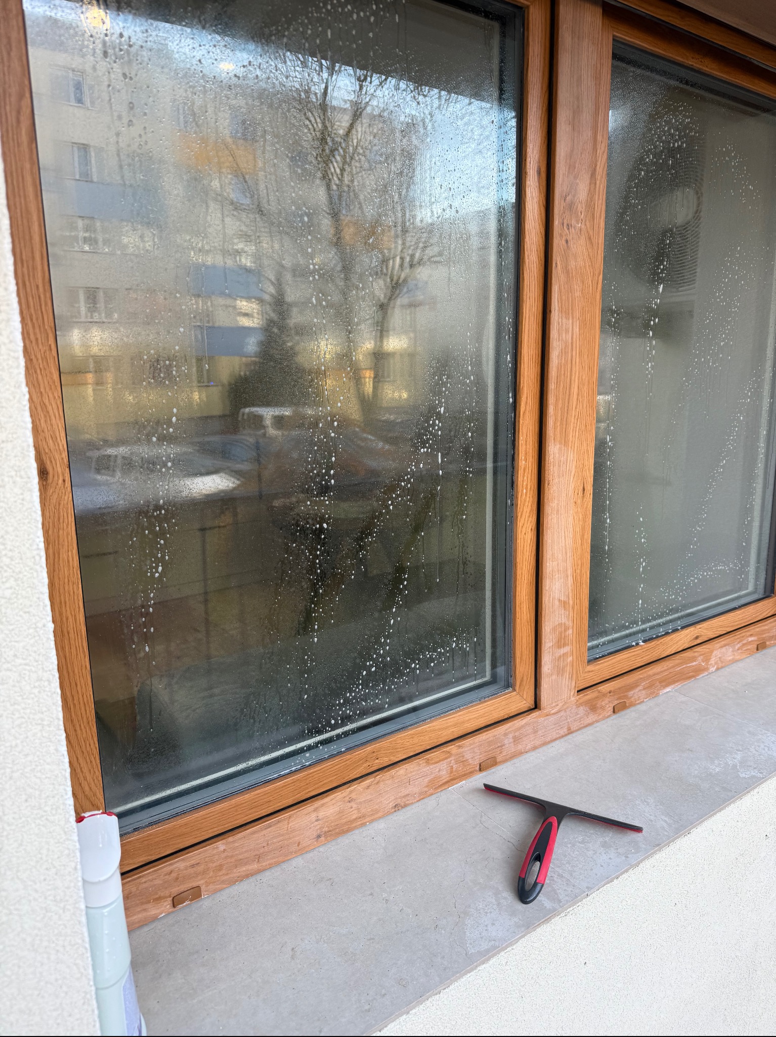 Brudne okno z drewnianą ramą, pokryte kroplami wody, obok leży ściągaczka. Widok na budynek mieszkalny za oknem. Częściowo umyte okno.